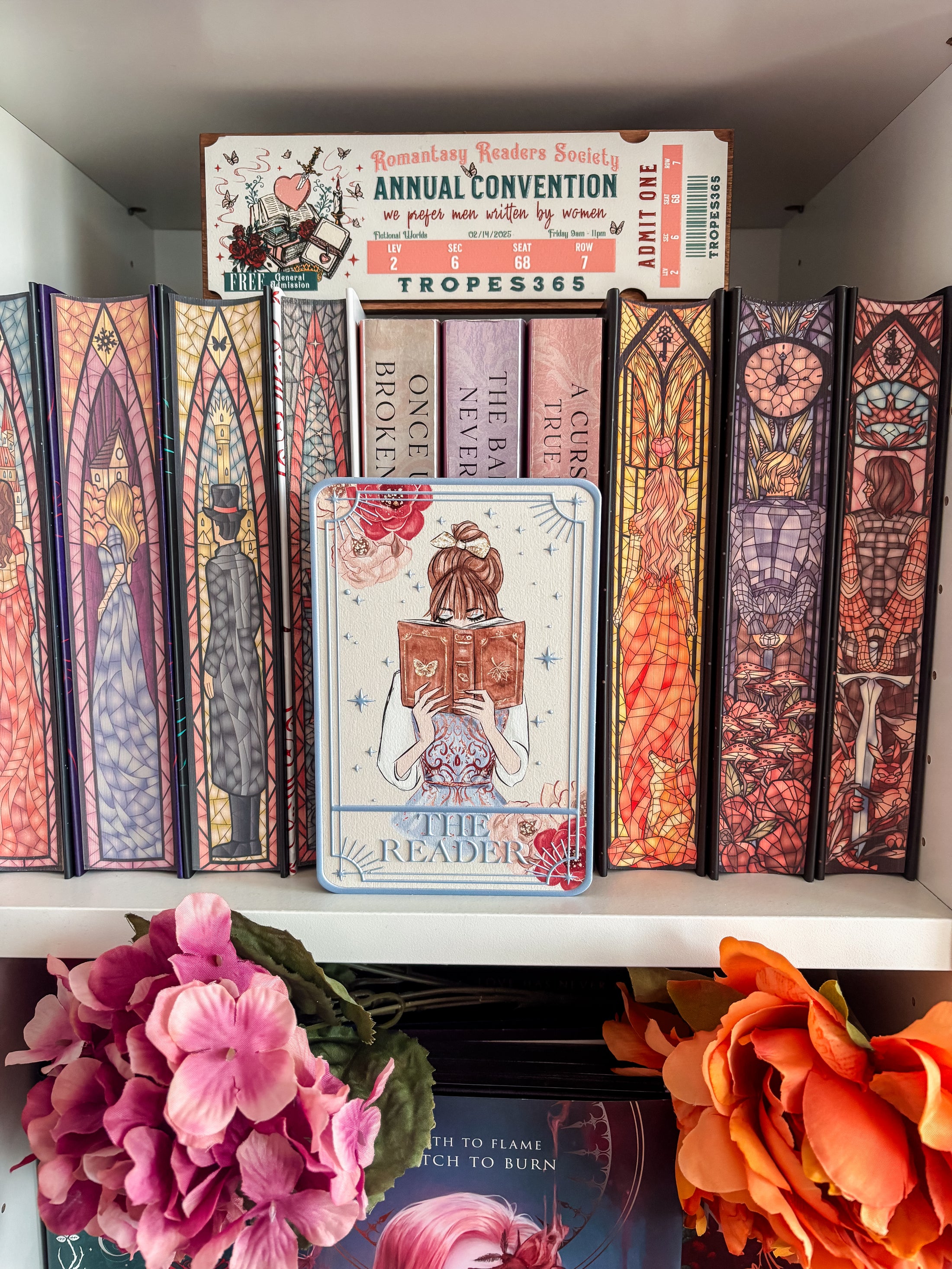 Tarot card title “The Reader” with a girl reading an open book in front of a bookshelf with flowers and other decorative items in the background. 