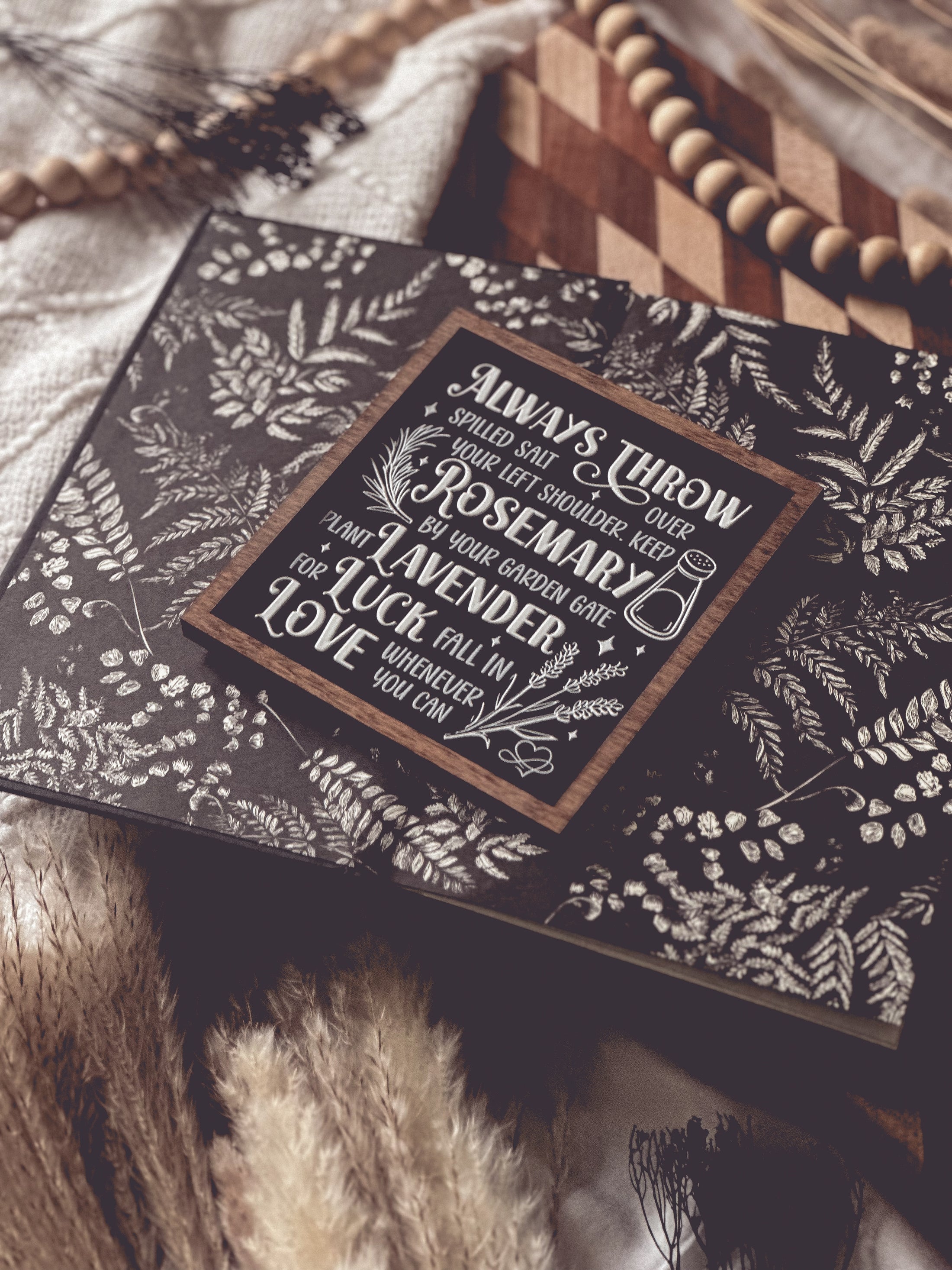 Black shelf sign with white text surrounded by a dark wood border. Graphics of a vial, lavender, rosemary and stars surround text that reads: “Always throw salt over your left shoulder. Keep rosemary by your garden gate. Plant lavender for luck. Fall in love whenever you can.” 