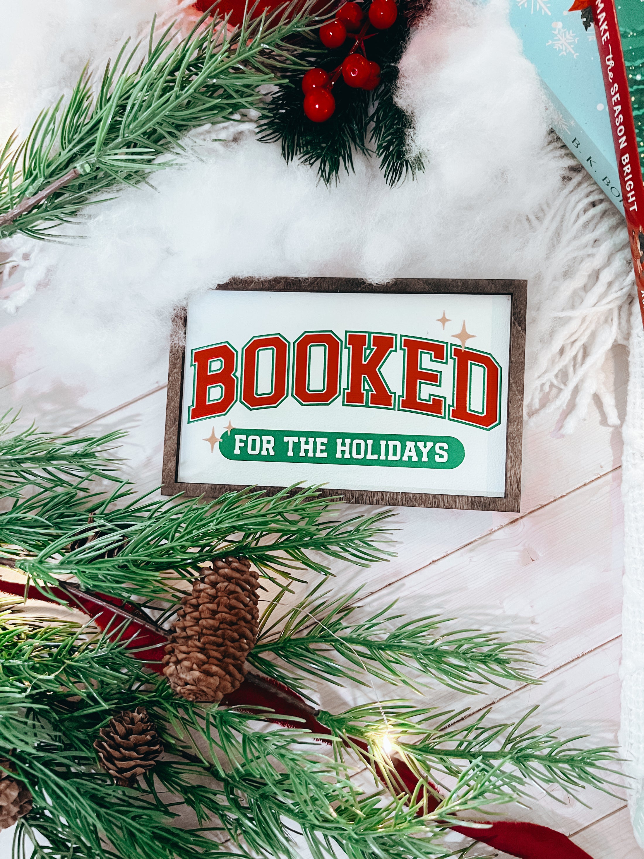 Decorative sign with a wooden frame that reads 'Booked for the Holidays' in red on a white background. © FireDrake Artistry 2025.