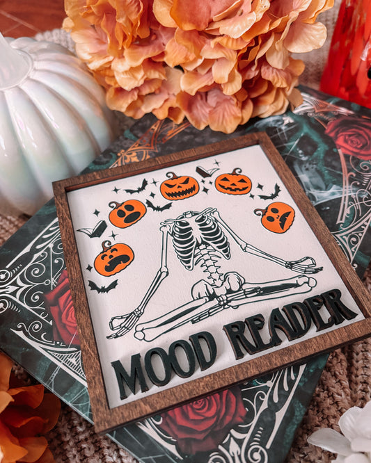 Mood Reader Sign. A square decorative sign made of wood with a wood-stained frame. UV printed skeleton in a zen position, surrounded by pumpkins with multiple faces, with the text 'MOOD READER' across the center. FireDrake Artistry 2025