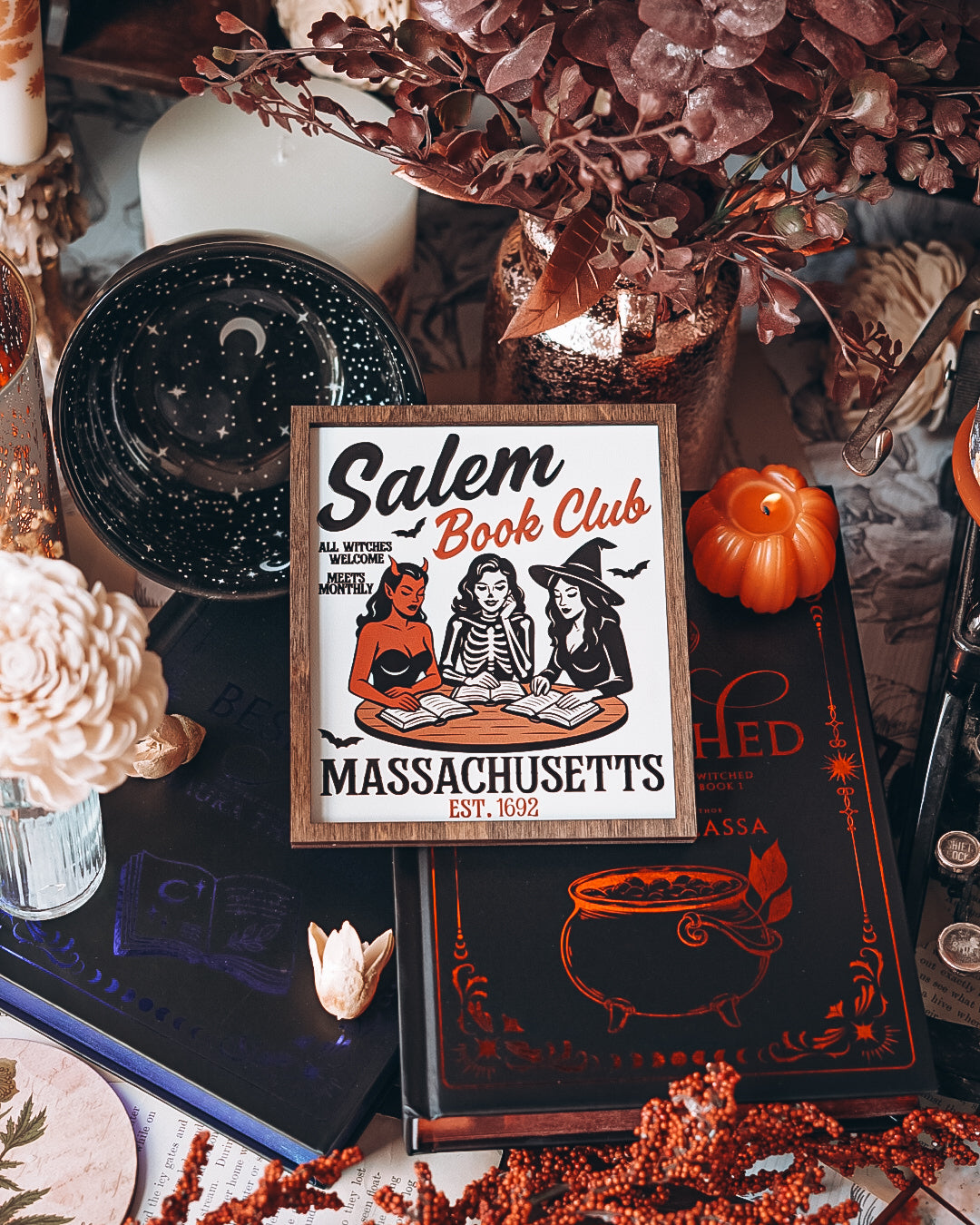 Salem Book Club Sign. White shelf sign with a wood grain border featuring a devil, witch, and skeleton women at a table reading. Top reads “SALEM BOOK CLUB” and the bottom reads “MASSACHUSETTS EST 1692” ©️Fire Drake Artistry 2025