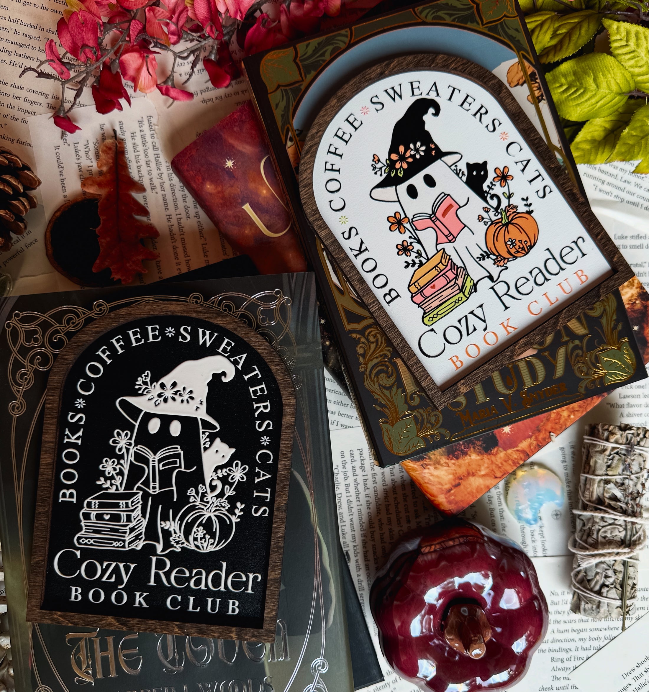 Cozy Reader Ghosty Sign. Two wooden, painted & UV printed signs. Left: black background with a dark wood border with a white design engraved. Right: white background with a dark wood border, colored endraved. Both feature a ghost reading a book and wearing a witch hat, surrounded by flowers and a pumpkin. Text above reads “Books. Coffee. Sweaters. Cats.” While the text below reads “Cozy Reader Book Club.” 