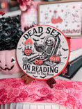 Load image into Gallery viewer, Dead Set on Reading Sign - Pink. White round sign with a skeleton lounging and reading a book surrounded by pumpkins. 
