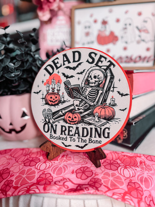 Dead Set on Reading Sign - Pink. White round sign with a skeleton lounging and reading a book surrounded by pumpkins. 