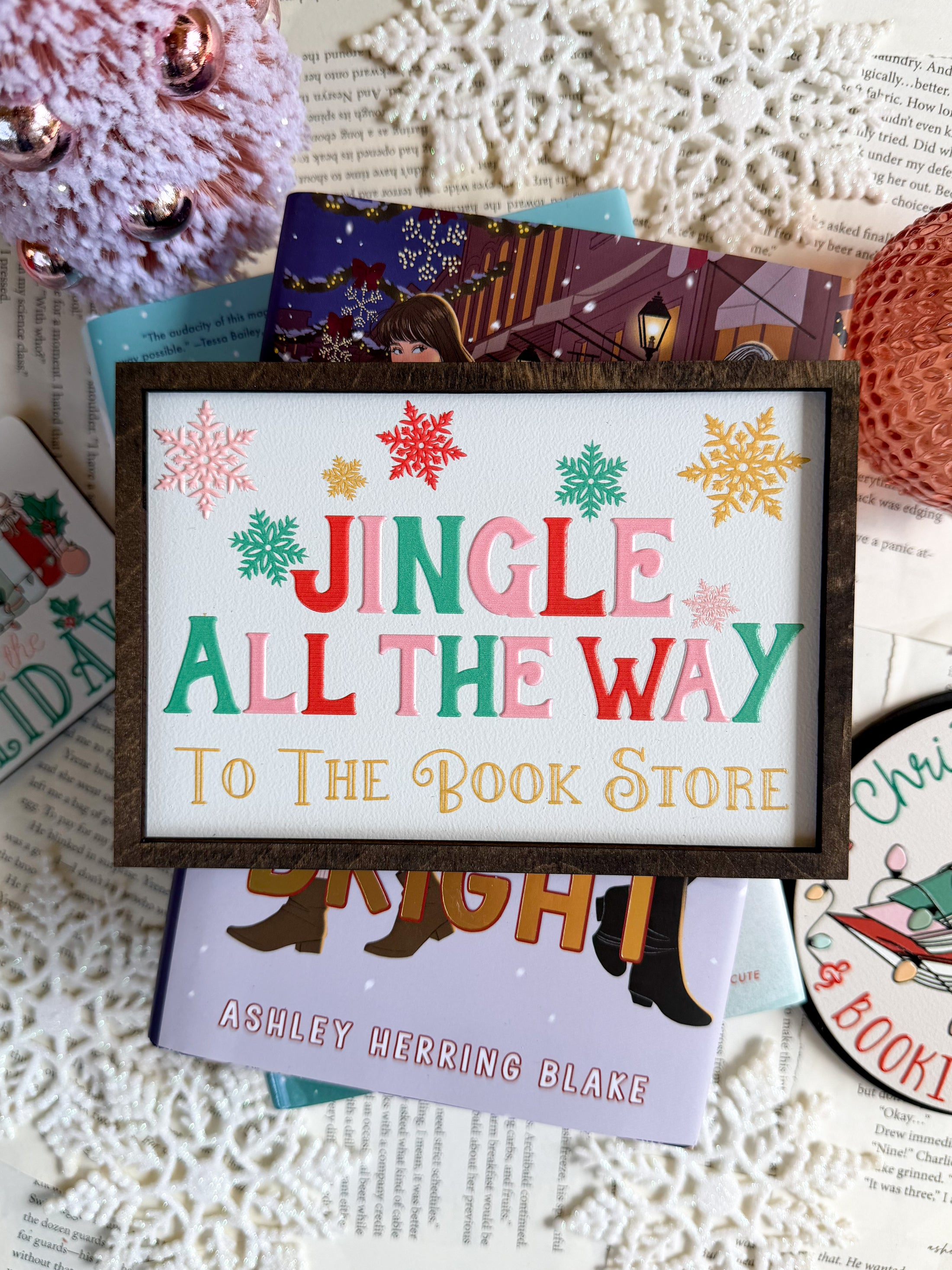 Decorative sign with a white background and woodgrain frame. Text reads 'Jingle All The Way To The Book Store' in Christmas colors, surrounded by snowflakes. © FireDrake Artistry 2025