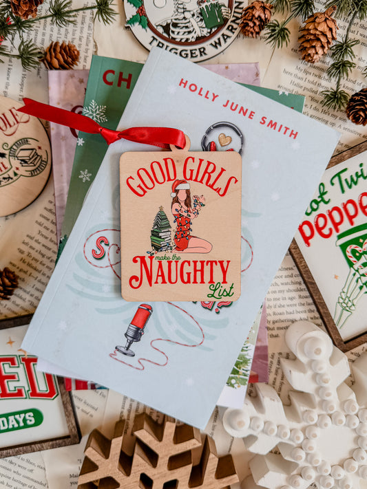 A rectangular wooden ornament featuring a Christmas tree of books next to a girl in a Santa hat tied up with Christmas lights surrounded by text that reads 'Good Girls Naughty List'. © FireDrake Artistry 2023