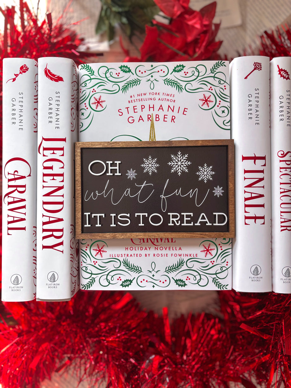 Wooden shelf sign with a black background and woodgrain frame. Featuring white text that reads "Oh what fun it is to read" and snowflakes. © FireDrake Artistry 2024.