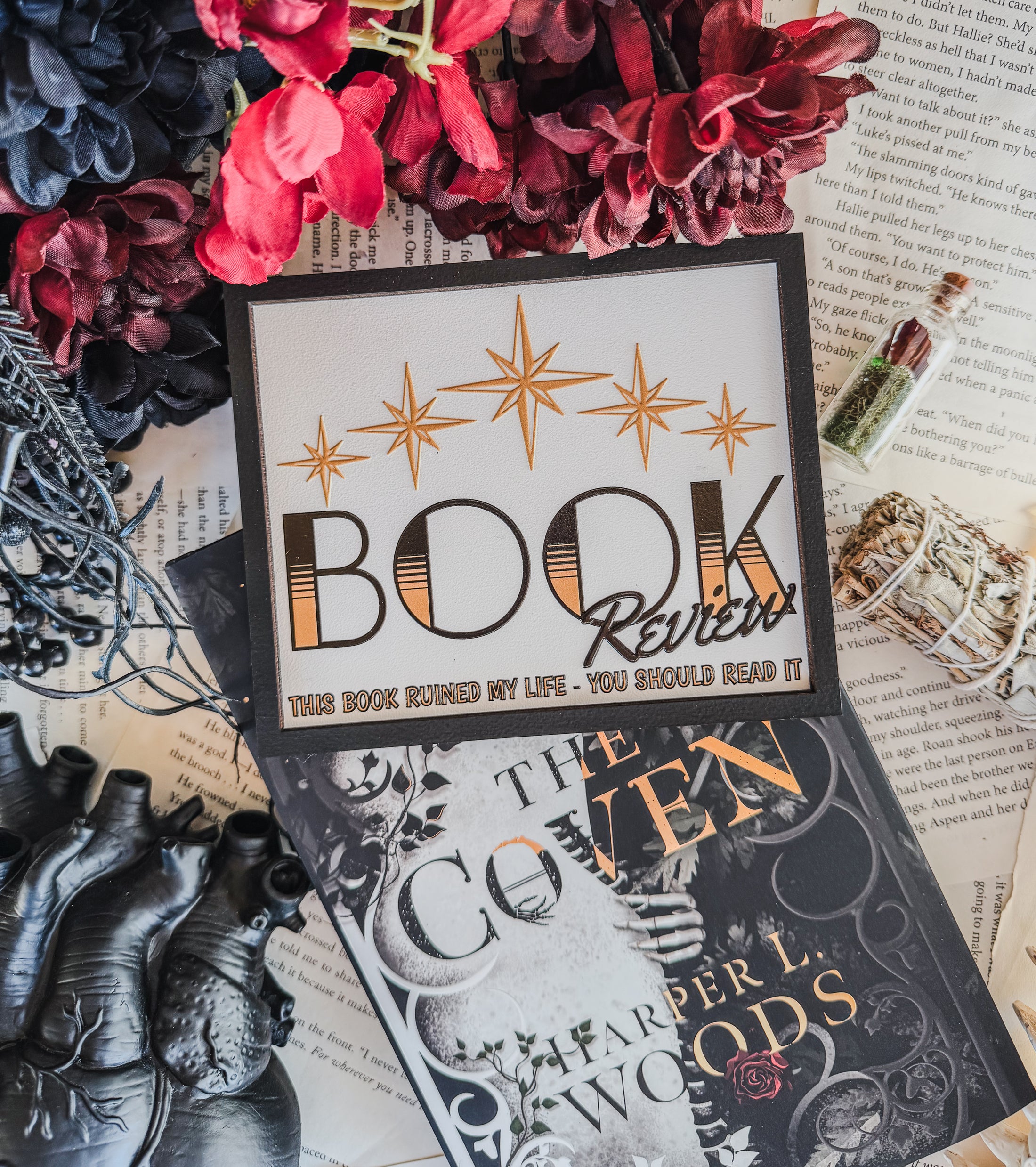 Wooden sign with 5 gold stars and the words “Book Review. This book ruined my life, you should read it” . Books, flowers, and book pages in the background. 