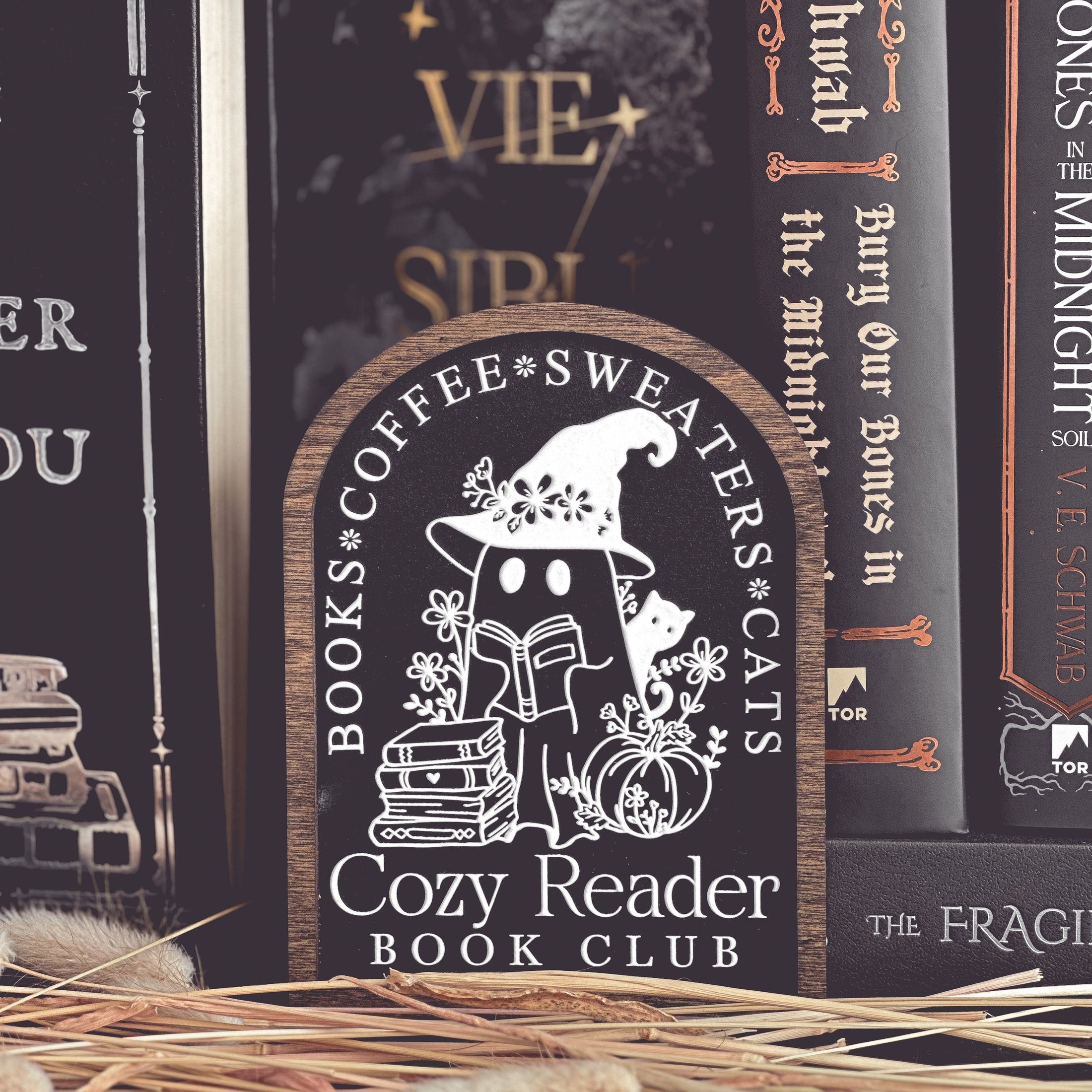 Cozy Reader Ghosty Sign. Black background with a dark wood border with a white design engraved. 
Featuring a ghost reading a book and wearing a witch hat, surrounded by flowers and a pumpkin. Text above reads “Books. Coffee. Sweaters. Cats.” While the text below reads “Cozy Reader Book Club.” 