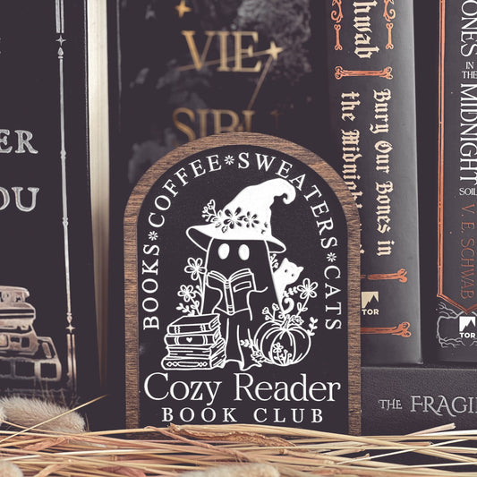 Cozy Reader Ghosty Sign. Black background with a dark wood border with a white design engraved. 
Featuring a ghost reading a book and wearing a witch hat, surrounded by flowers and a pumpkin. Text above reads “Books. Coffee. Sweaters. Cats.” While the text below reads “Cozy Reader Book Club.” 