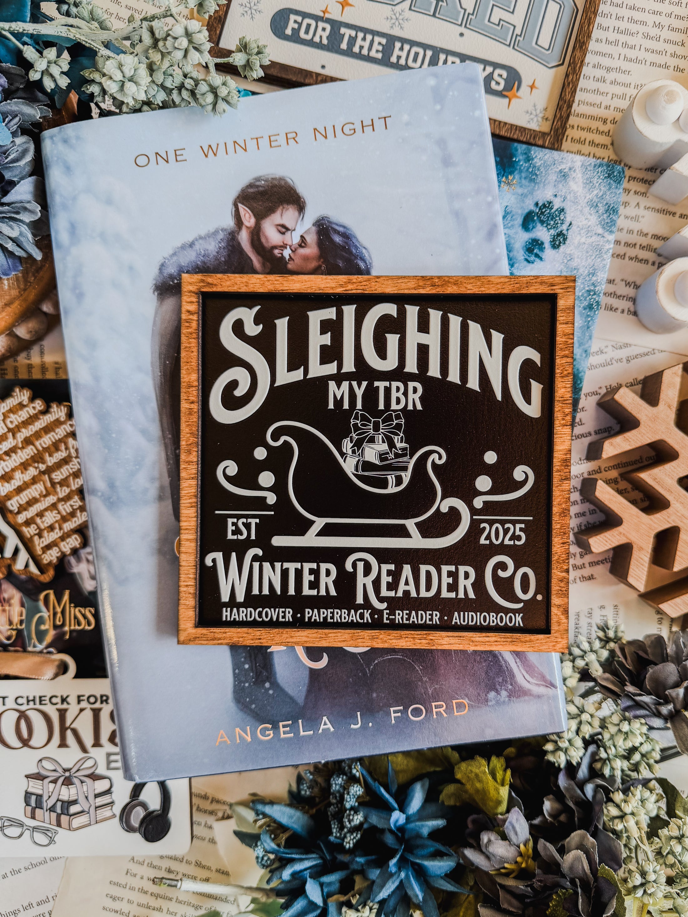 Wooden shelf sign with a black background and woodgrain frame.  A white sleigh with presents, surrounded by white text reading  'Sleighing My TBR' and "Winter Reader Co. EST 2025." © FireDrake Artistry 2025