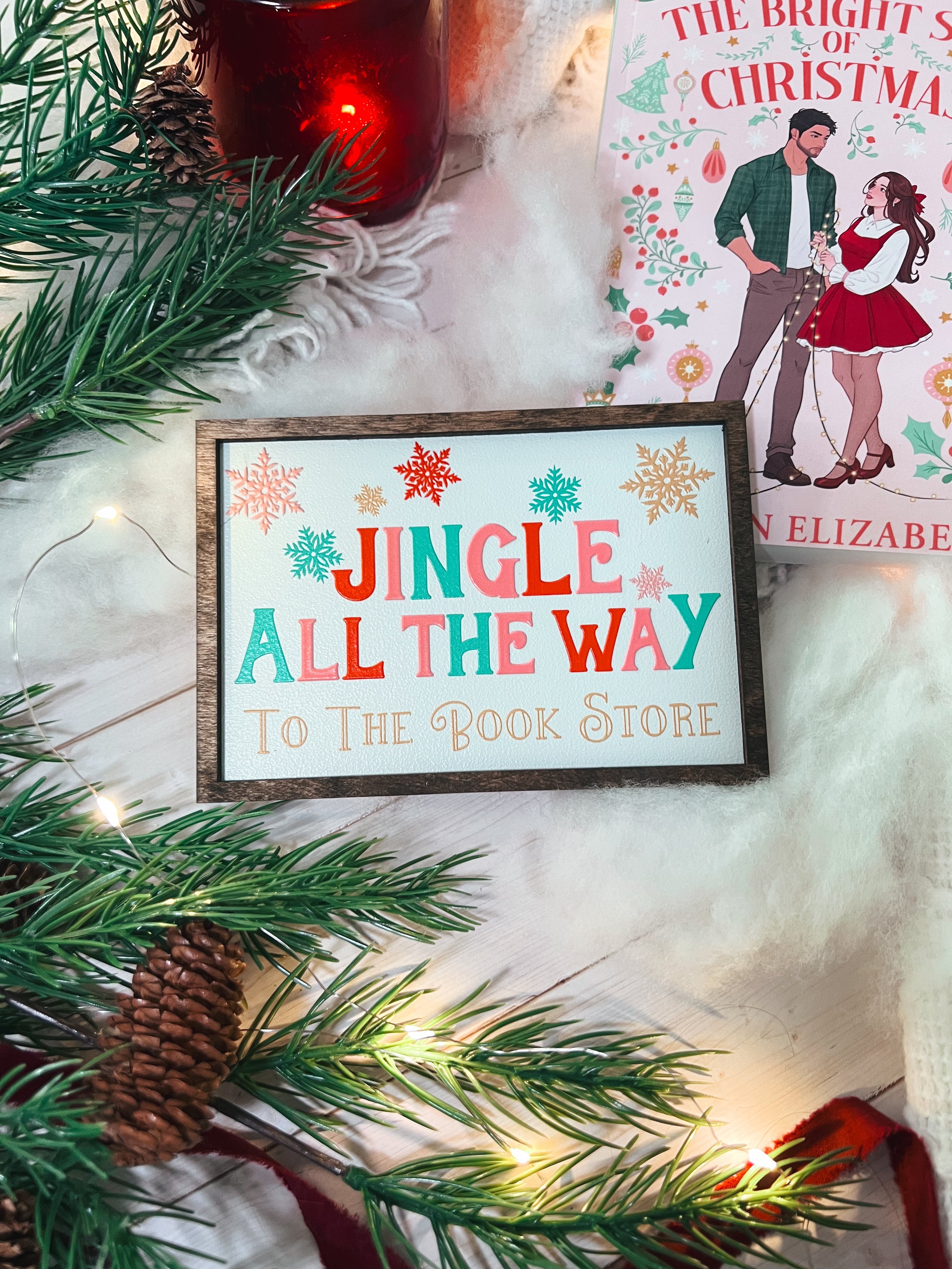 Decorative sign with a white background and woodgrain frame. Text reads 'Jingle All The Way To The Book Store' in Christmas colors, surrounded by snowflakes. © FireDrake Artistry 2025