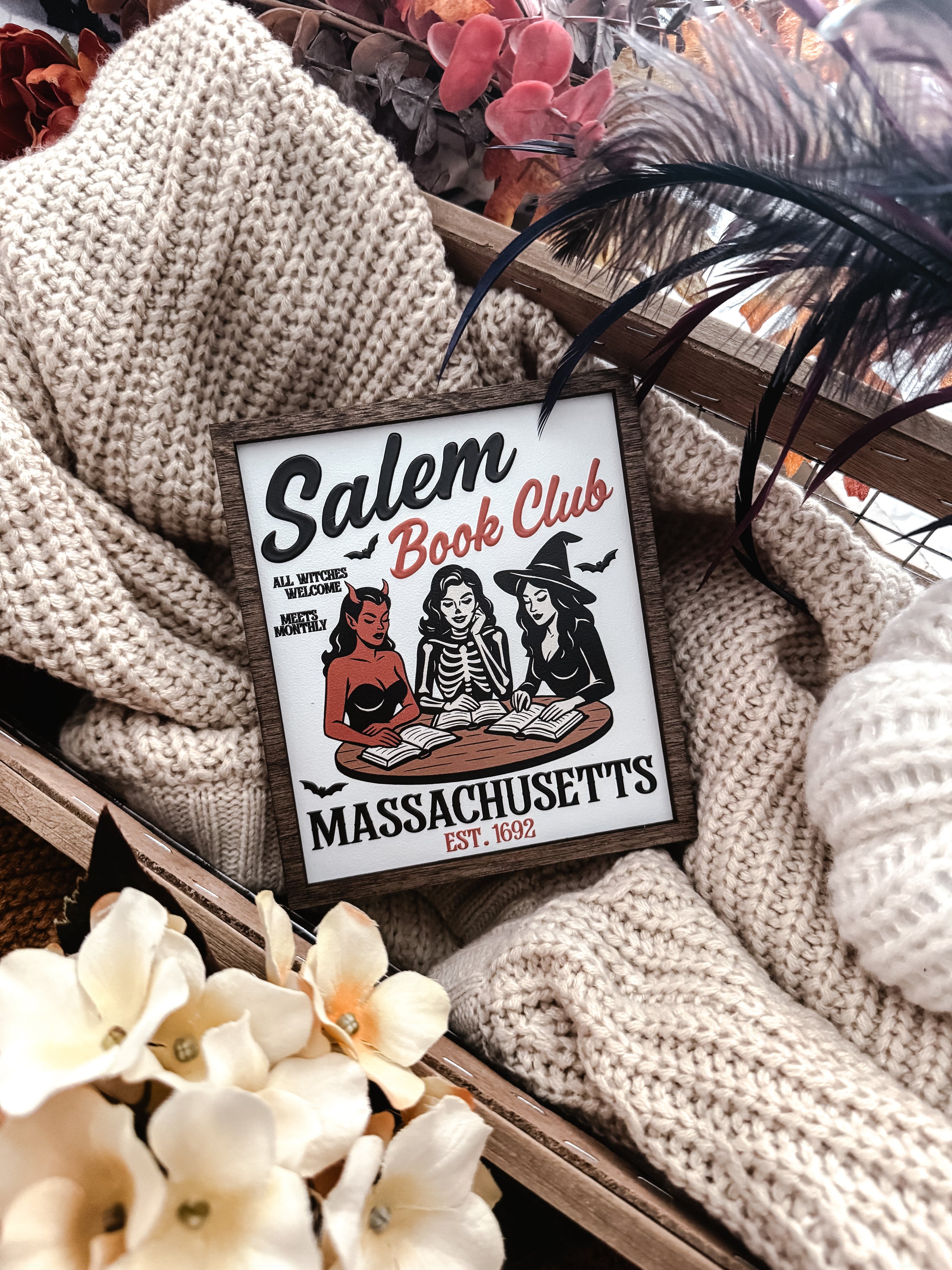 Salem Book Club Sign. White shelf sign with a wood grain border featuring a devil, witch, and skeleton women at a table reading. Top reads “SALEM BOOK CLUB” and the bottom reads “MASSACHUSETTS EST 1692” ©️Fire Drake Artistry 2025