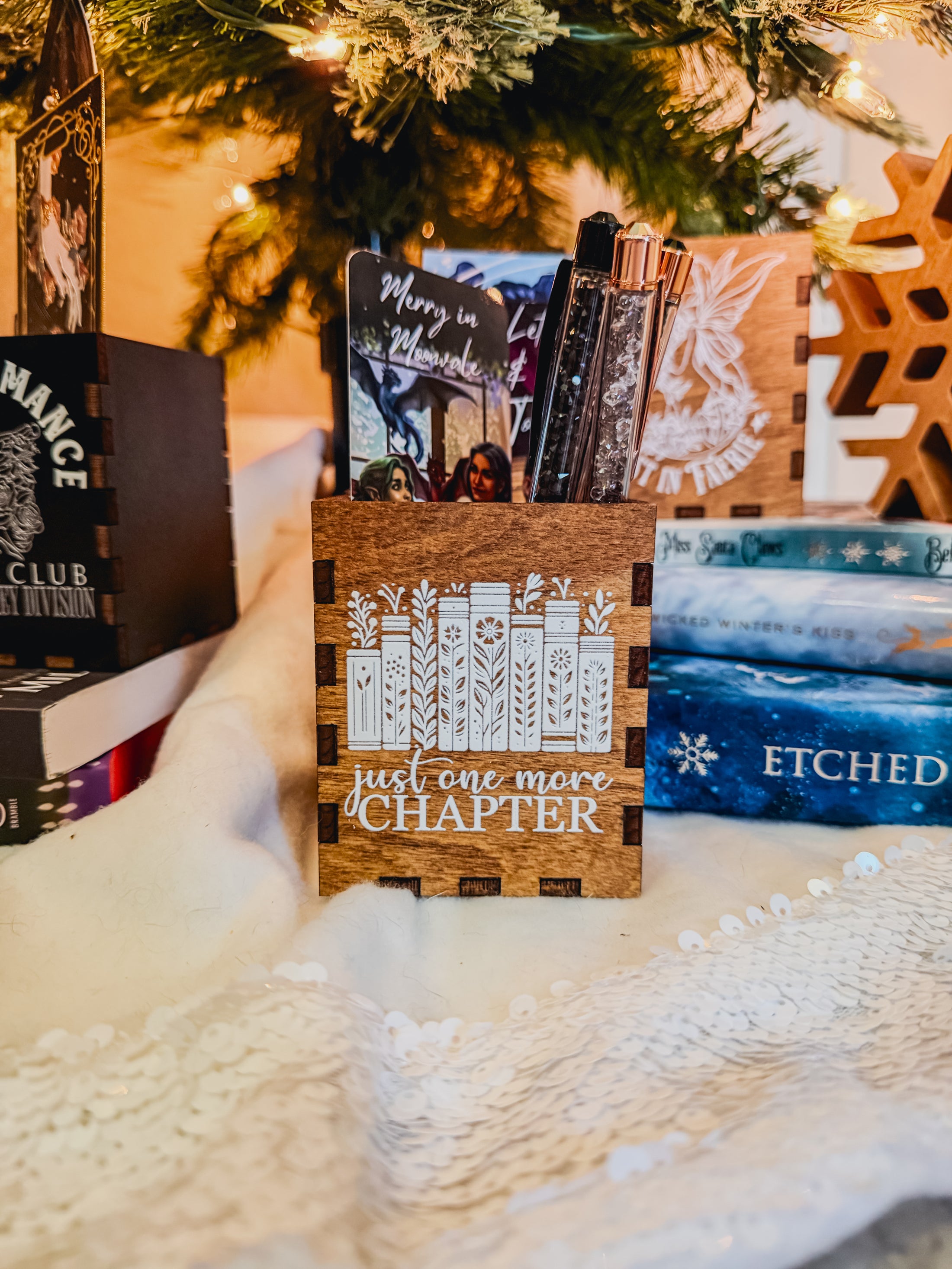 Wooden box with printed design with 'Just one more chapter' text and a book stack with flowers, against a physical books and Christmas tree background.