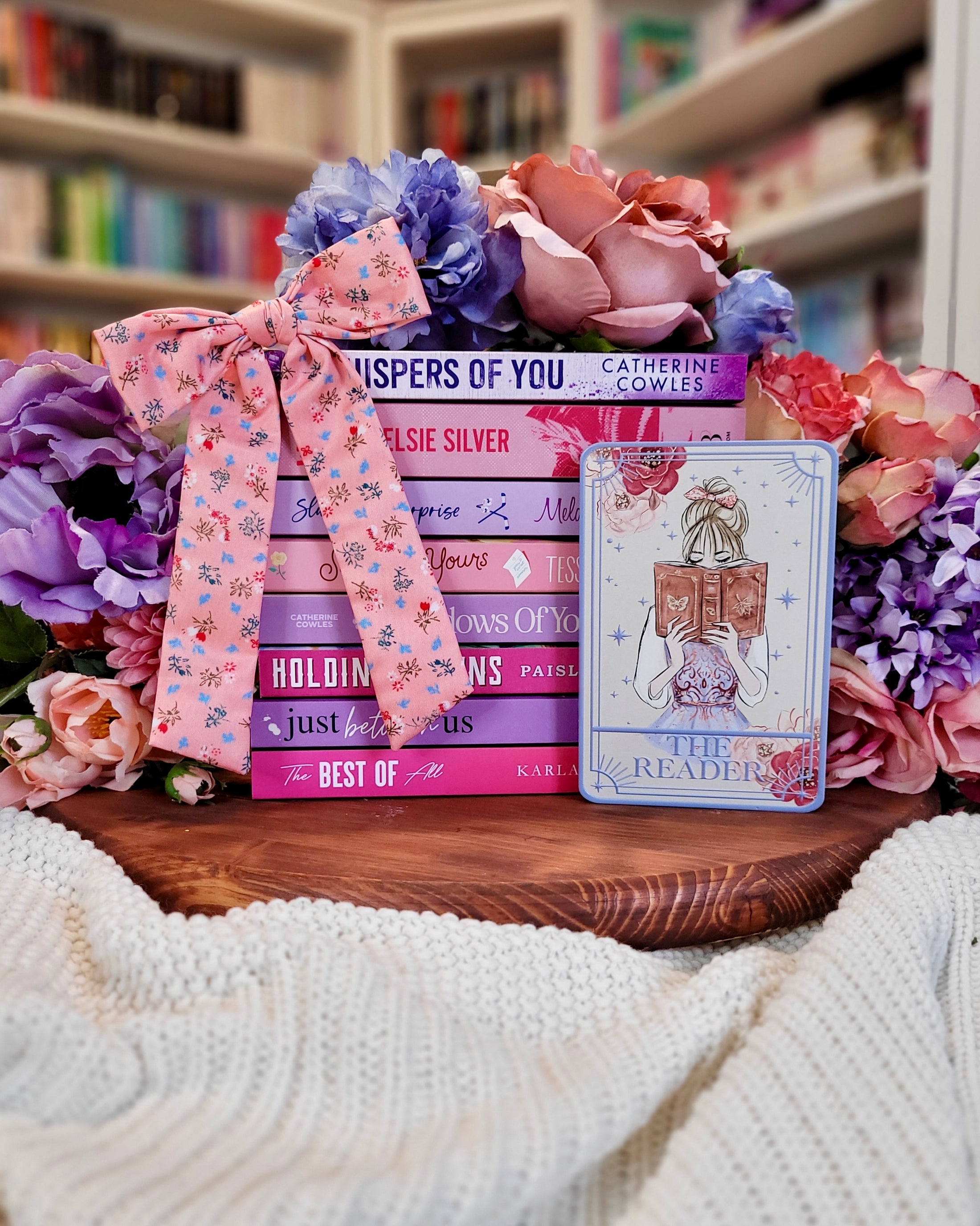 Tarot card title “The Reader” with a girl reading an open book in front of  a bookstack with flowers and other decorative items in the background. 