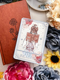Load image into Gallery viewer, Tarot card titled 'The Reader' surrounded by flowers and a notebook on a wooden surface
