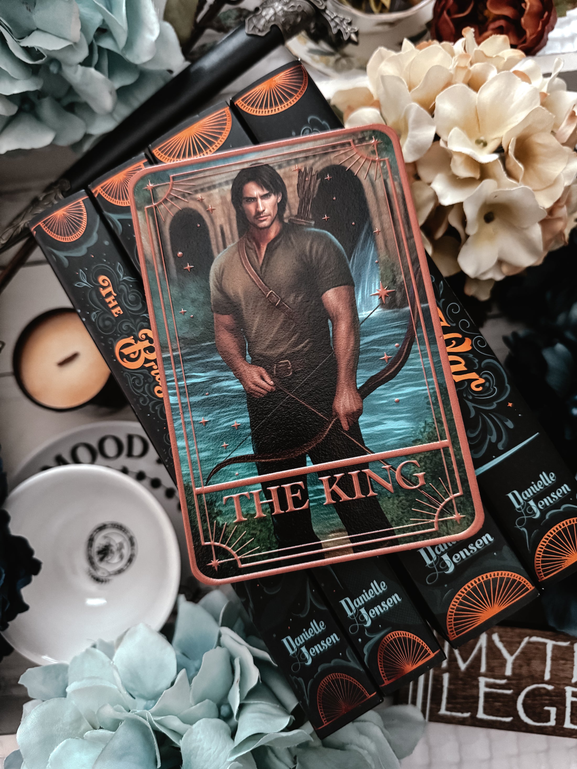 Tarot card featuring 'The King' won top of a star of books with flowers and decorative elements on a table in the background. 