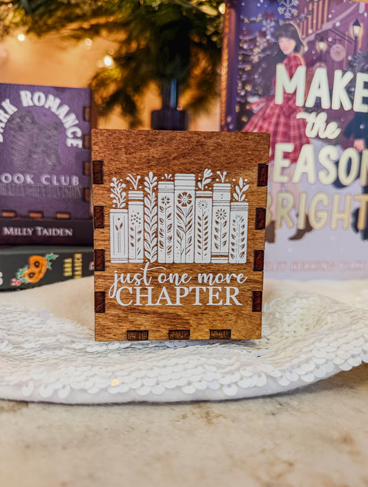 Wooden box with printed design with 'Just one more chapter' text and a book stack with flowers, against a physical books and Christmas tree background.