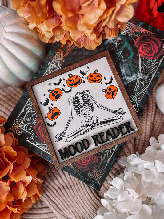 Mood Reader Sign. A square decorative sign made of wood with a wood-stained frame. UV printed skeleton in a zen position, surrounded by pumpkins with multiple faces, with the text 'MOOD READER' across the center. FireDrake Artistry 2025