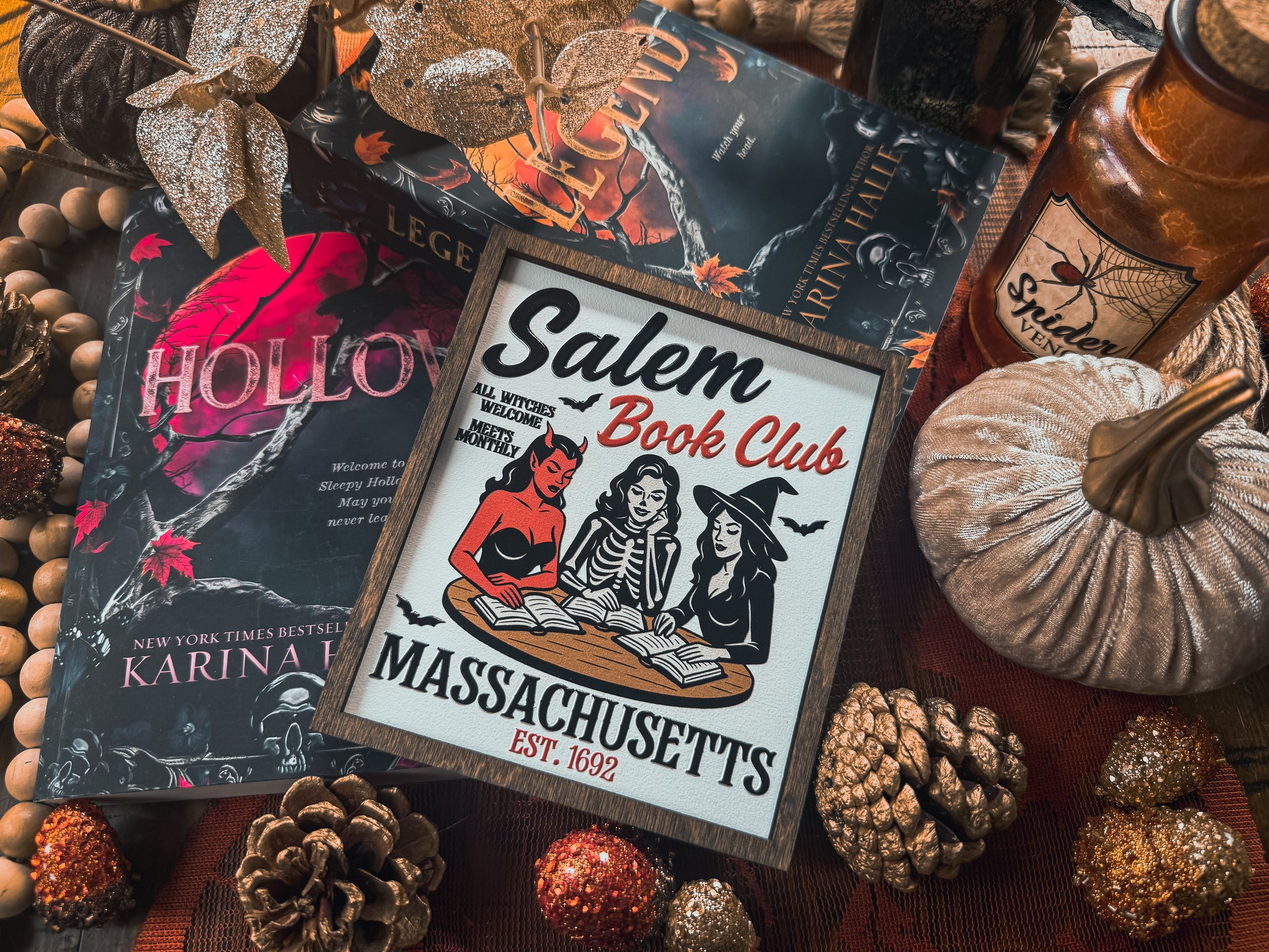 Salem Book Club Sign. White shelf sign with a wood grain border featuring a devil, witch, and skeleton women at a table reading. Top reads “SALEM BOOK CLUB” and the bottom reads “MASSACHUSETTS EST 1692” ©️Fire Drake Artistry 2025