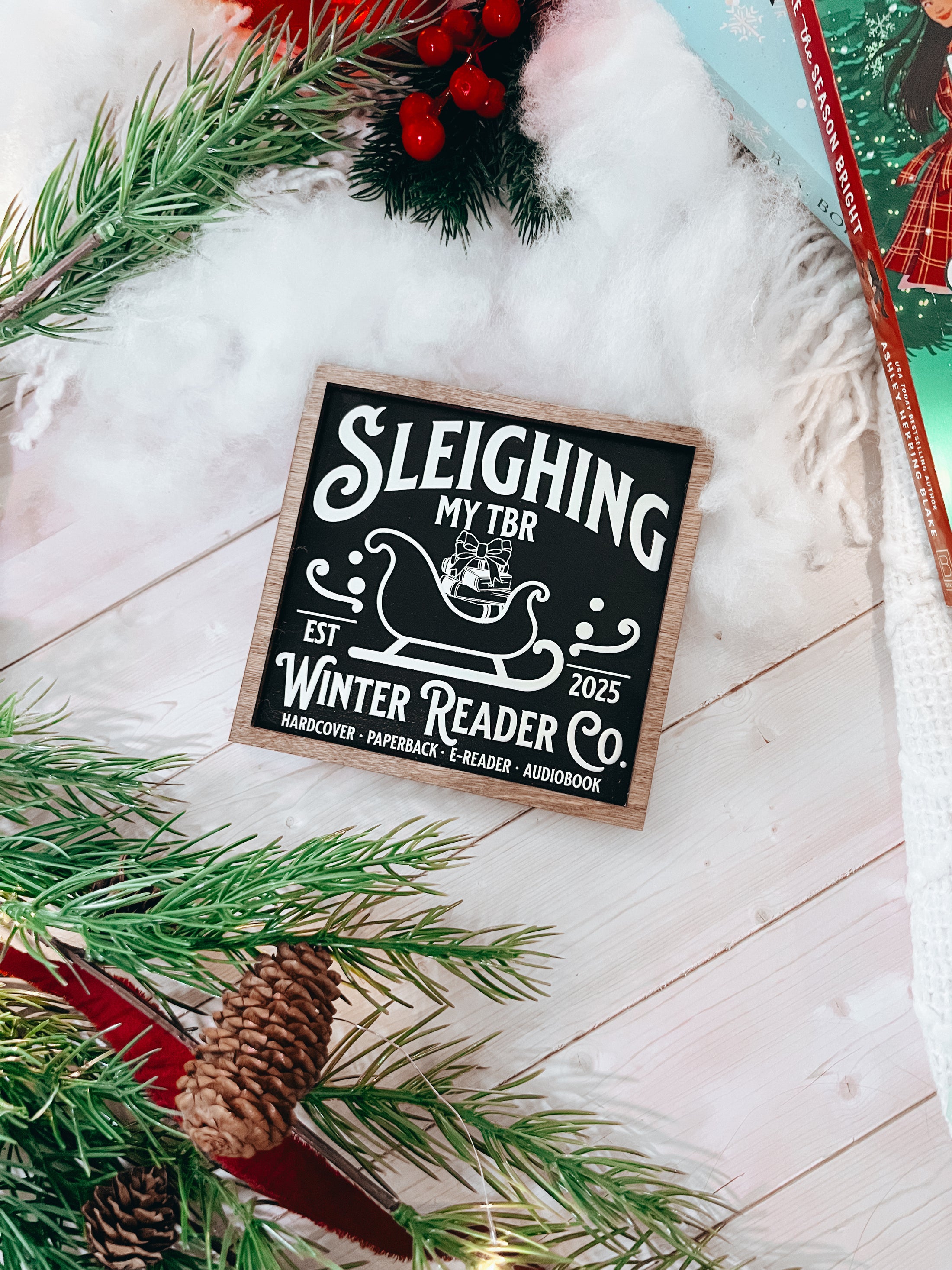 Wooden shelf sign with a black background and woodgrain frame.  A white sleigh with presents, surrounded by white text reading  'Sleighing My TBR' and "Winter Reader Co. EST 2025." © FireDrake Artistry 2025