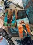 Load image into Gallery viewer, Two tarot cards, 'The King' and 'The Queen', on a surface with decorative elements of books and flowers. 
