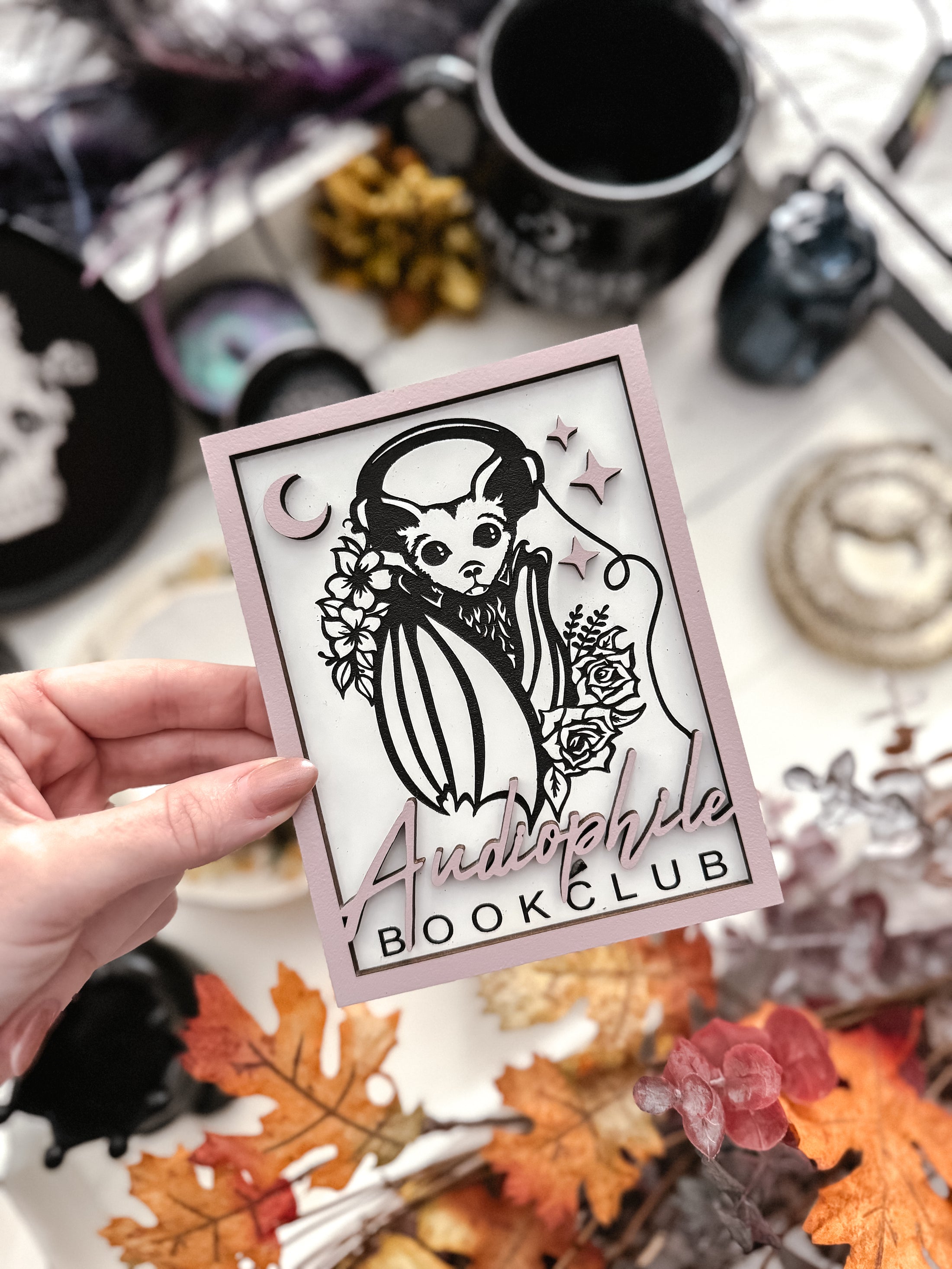 Audiophile Book Club Sign. White wooden sign with a lilac border. Bat design with headphones surrounded by flowers, with words below that read "Audiophile Book Club". 2023 FireDrake Artistry®