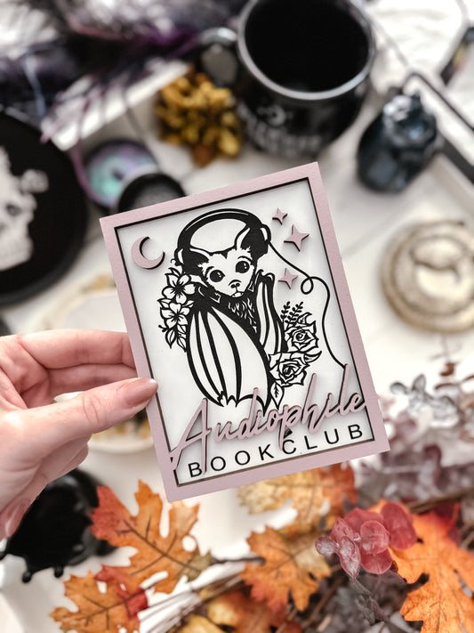 Audiophile Book Club Sign. White wooden sign with a lilac border. Bat design with headphones surrounded by flowers, with words below that read "Audiophile Book Club". 2023 FireDrake Artistry®