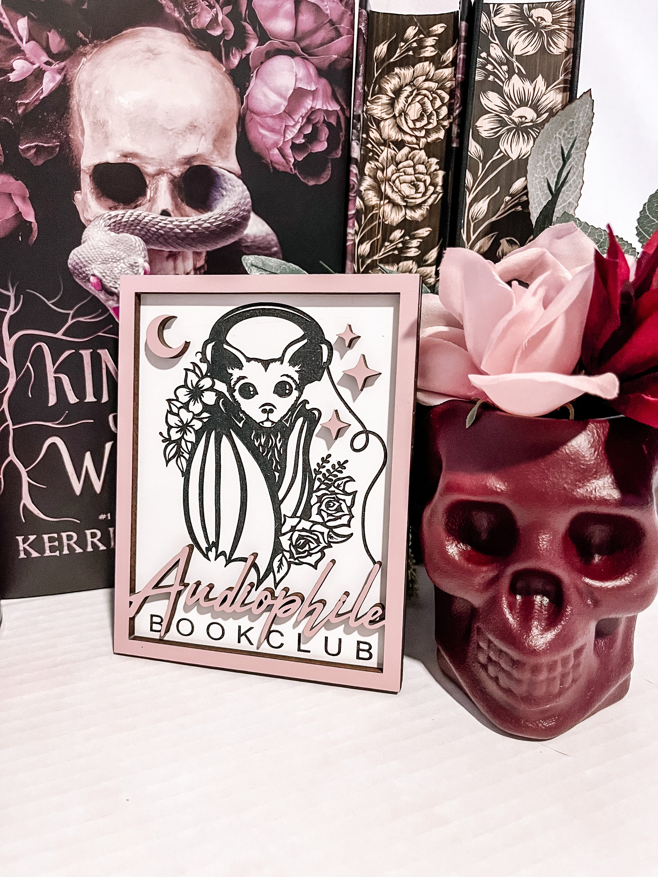 Audiophile Book Club Sign. White wooden sign with a lilac border. Bat design with headphones surrounded by flowers, with words below that read "Audiophile Book Club". 2023 FireDrake Artistry®