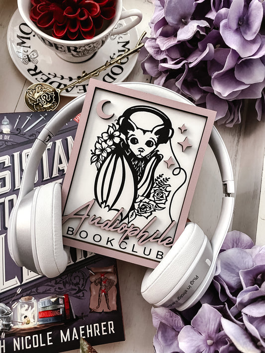 Audiophile Book Club Sign. White wooden sign with a lilac border. Bat design with headphones surrounded by flowers, with words below that read "Audiophile Book Club". 2023 FireDrake Artistry®