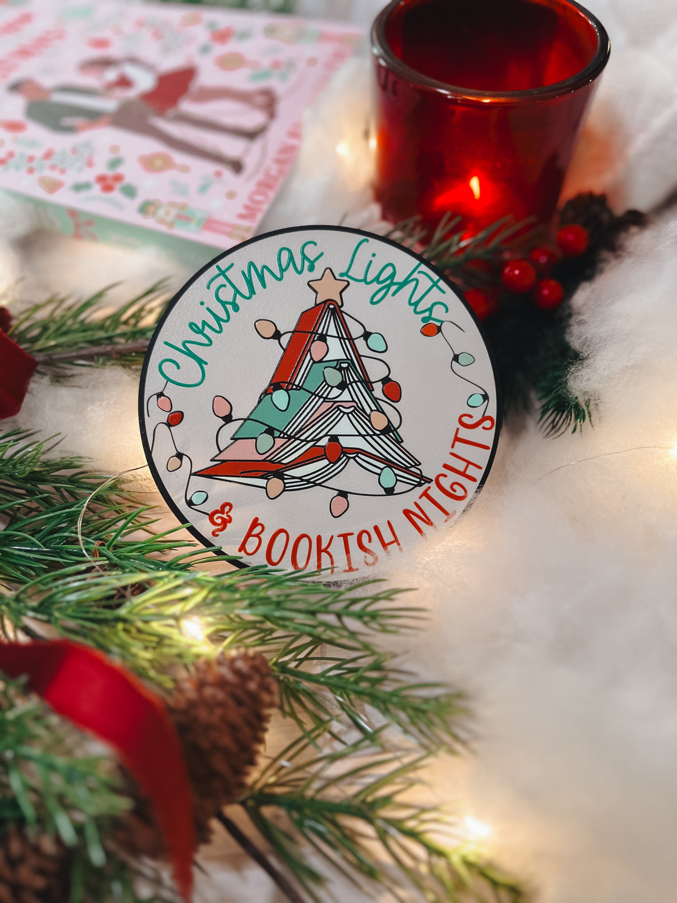 Round shelf sign with a white background. Image of books stacked into a Christmas tree surrounded by lights. The surrounding text reads "Christmas Lights" above in green, with "& Bookish Nights" below in red. © FireDrake Artistry 2025