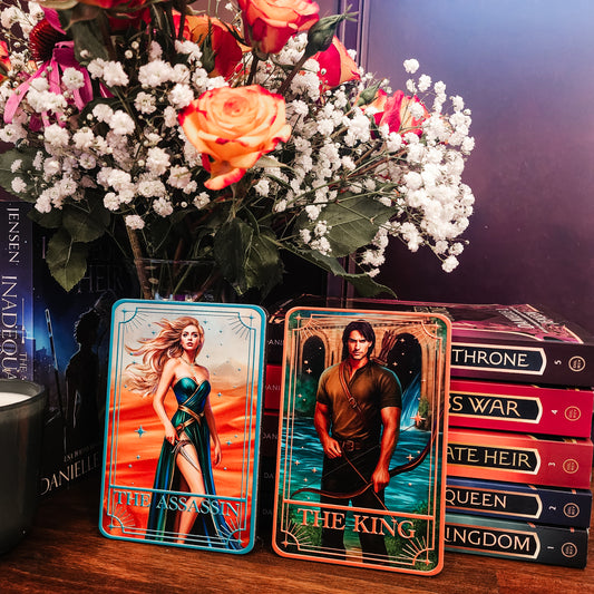 Two tarot cards titled 'The Assassin' and 'The King' with a bouquet of flowers and books in the background.