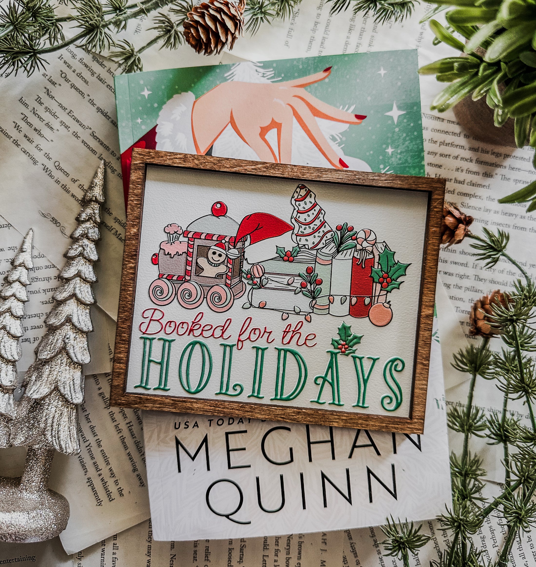 Wooden sign with a white background and woodgrain frame. Graphic features a gingerbread train pulling a stack of books with other Christmas items with text below that reads "Booked for the Holidays". © FireDrake Artistry 2024