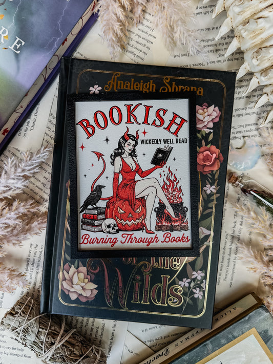 White shelf sign with a black border featuring a graphic of a woman in a devil costume reading a book and sitting on a pumpkin. Surrounding text reads “Bookish: burning through books”