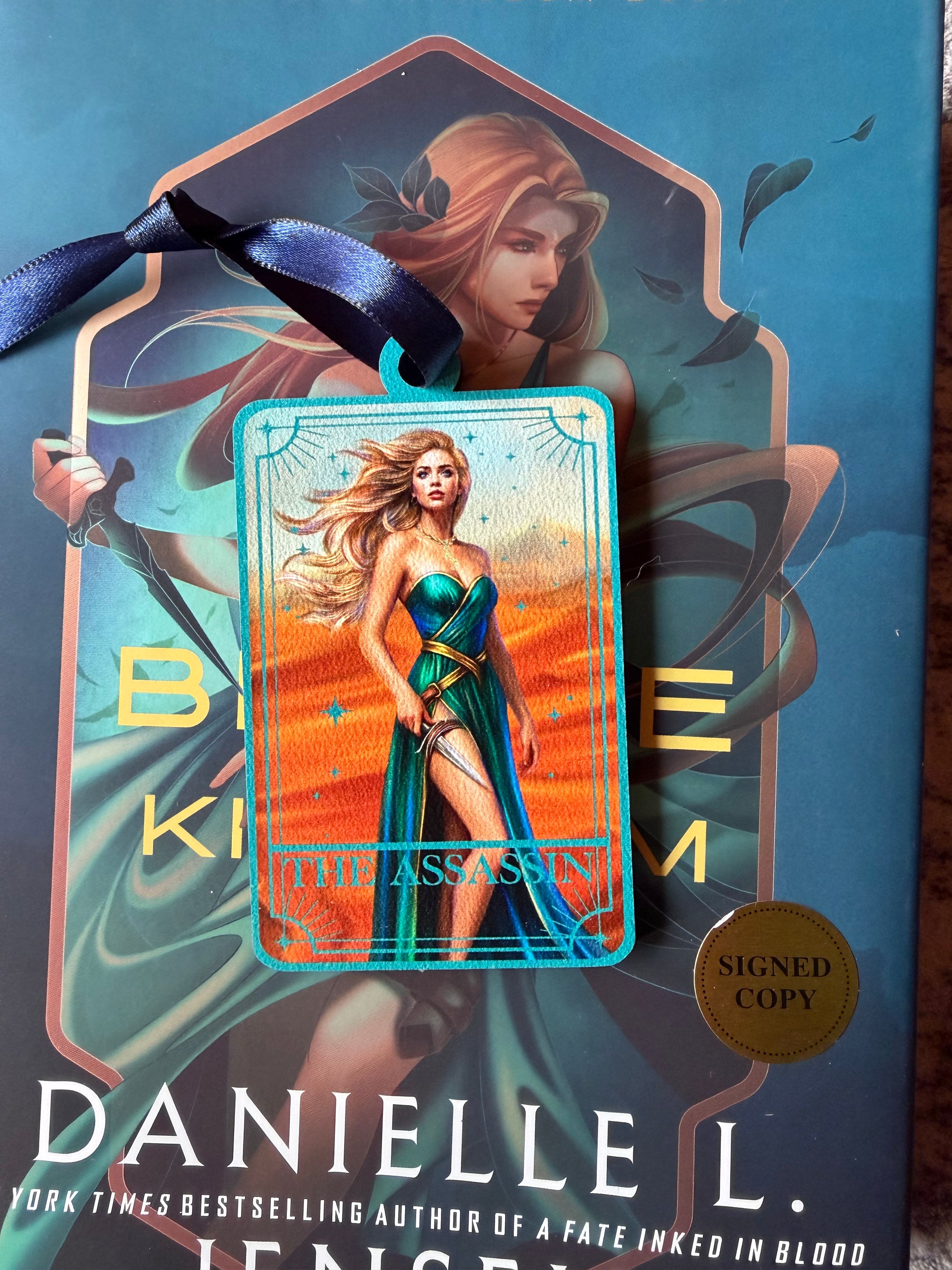 Tarot Card ornament titled "The Assassin" with  blue ribbon and book underneath.