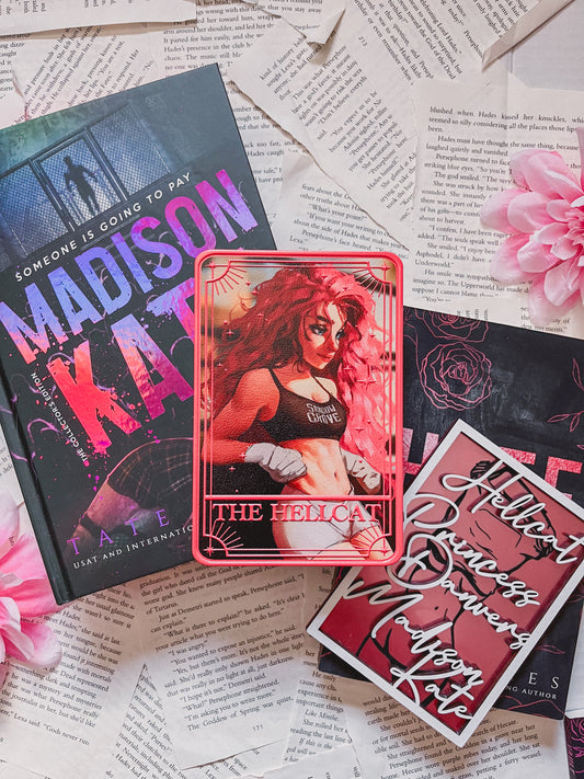 A pink tarot card labeled "The Hellcat" on top of a book titled Madison Kate by Tate James with a shelf sign, books, flowers, and book pages in the background. 