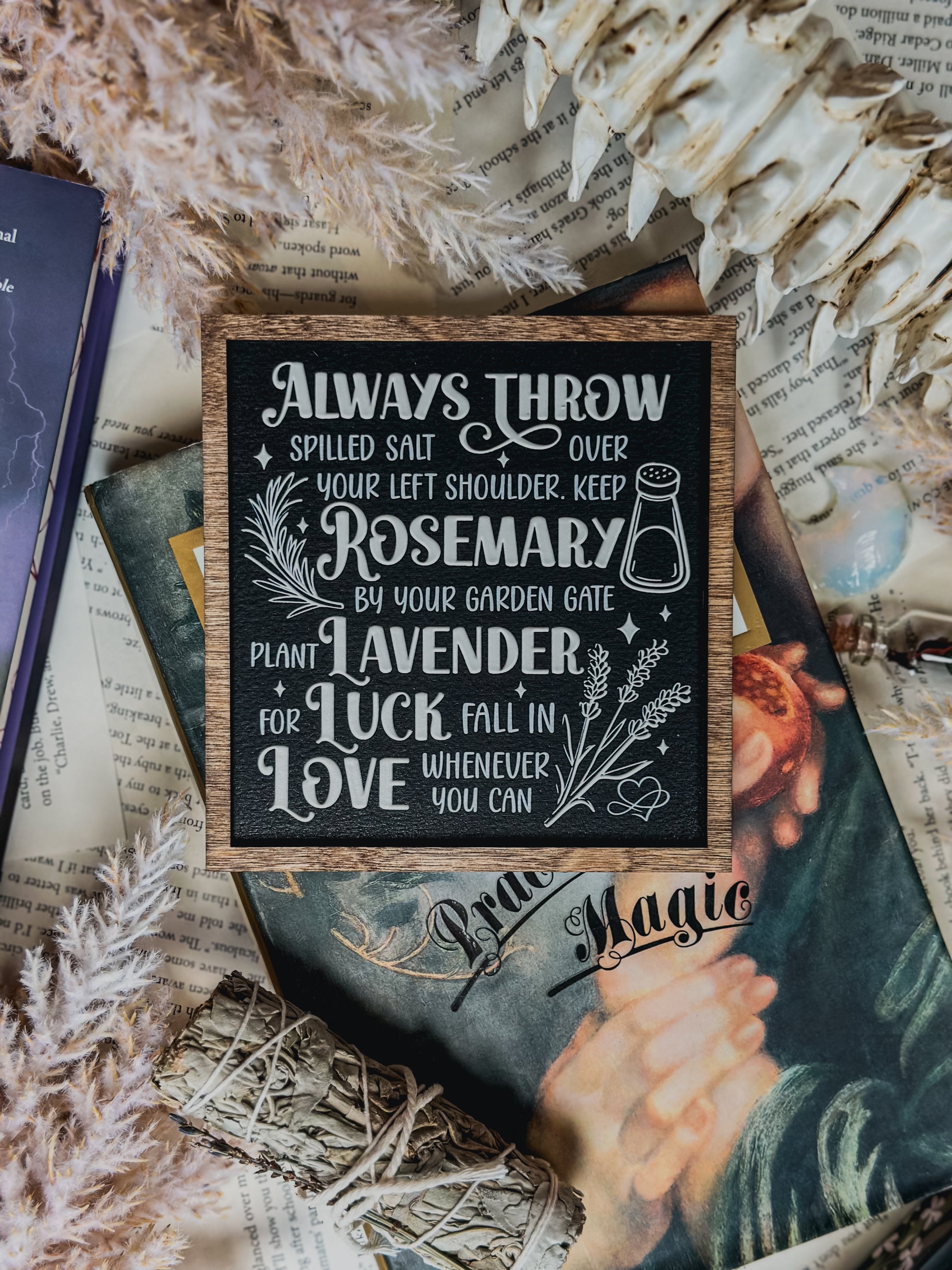 Black shelf sign with white text surrounded by a dark wood border. Graphics of a vial, lavender, rosemary and stars surround text that reads: “Always throw salt over your left shoulder. Keep rosemary by your garden gate. Plant lavender for luck. Fall in love whenever you can”. 