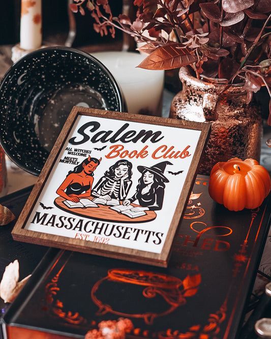 Salem Book Club Sign. White shelf sign with a wood grain border featuring a devil, witch, and skeleton women at a table reading. Top reads “SALEM BOOK CLUB” and the bottom reads “MASSACHUSETTS EST 1692” ©️Fire Drake Artistry 2025