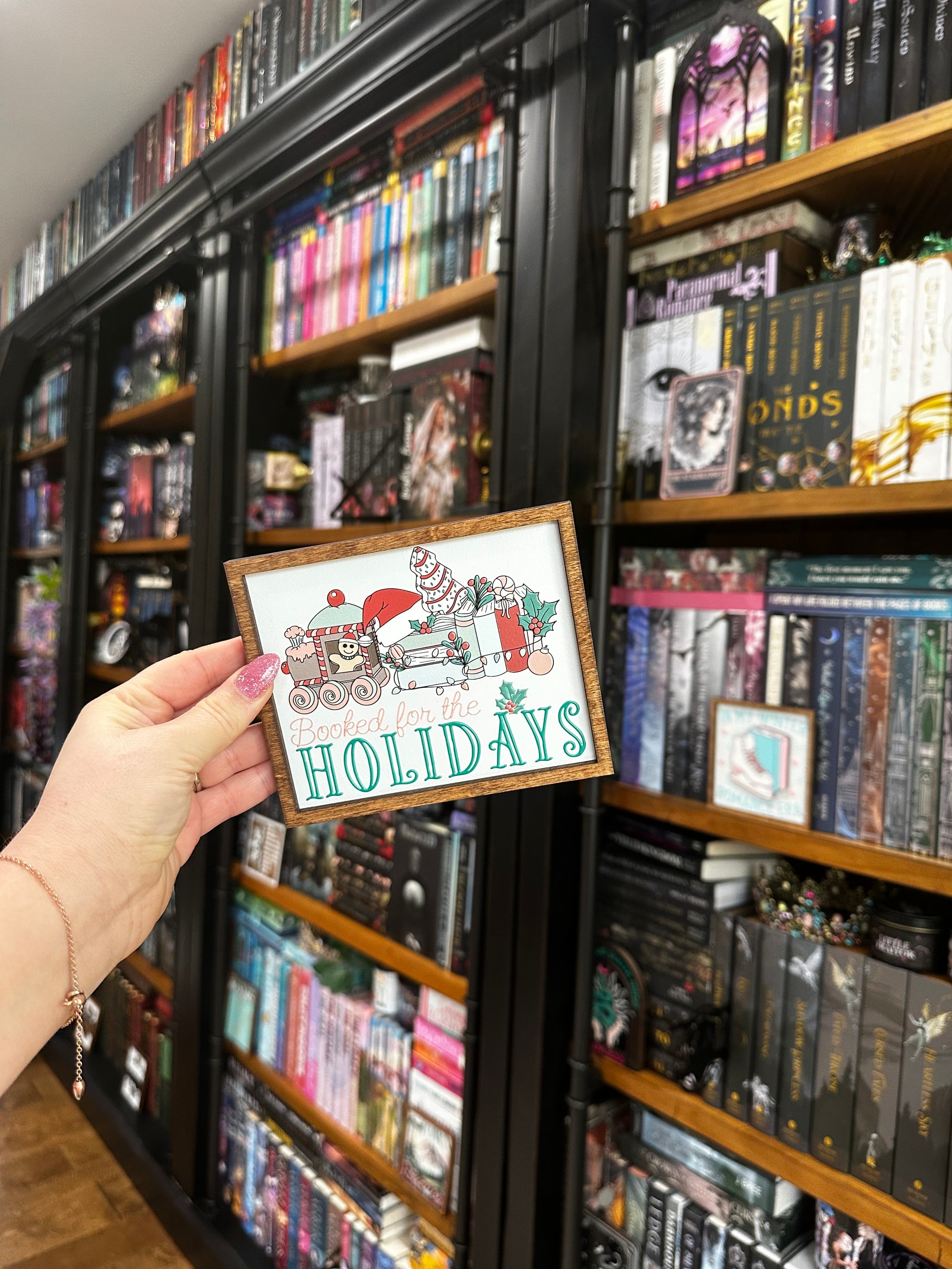 Wooden sign with a white background and woodgrain frame. Graphic features a gingerbread train pulling a stack of books with other Christmas items with text below that reads "Booked for the Holidays". © FireDrake Artistry 2024