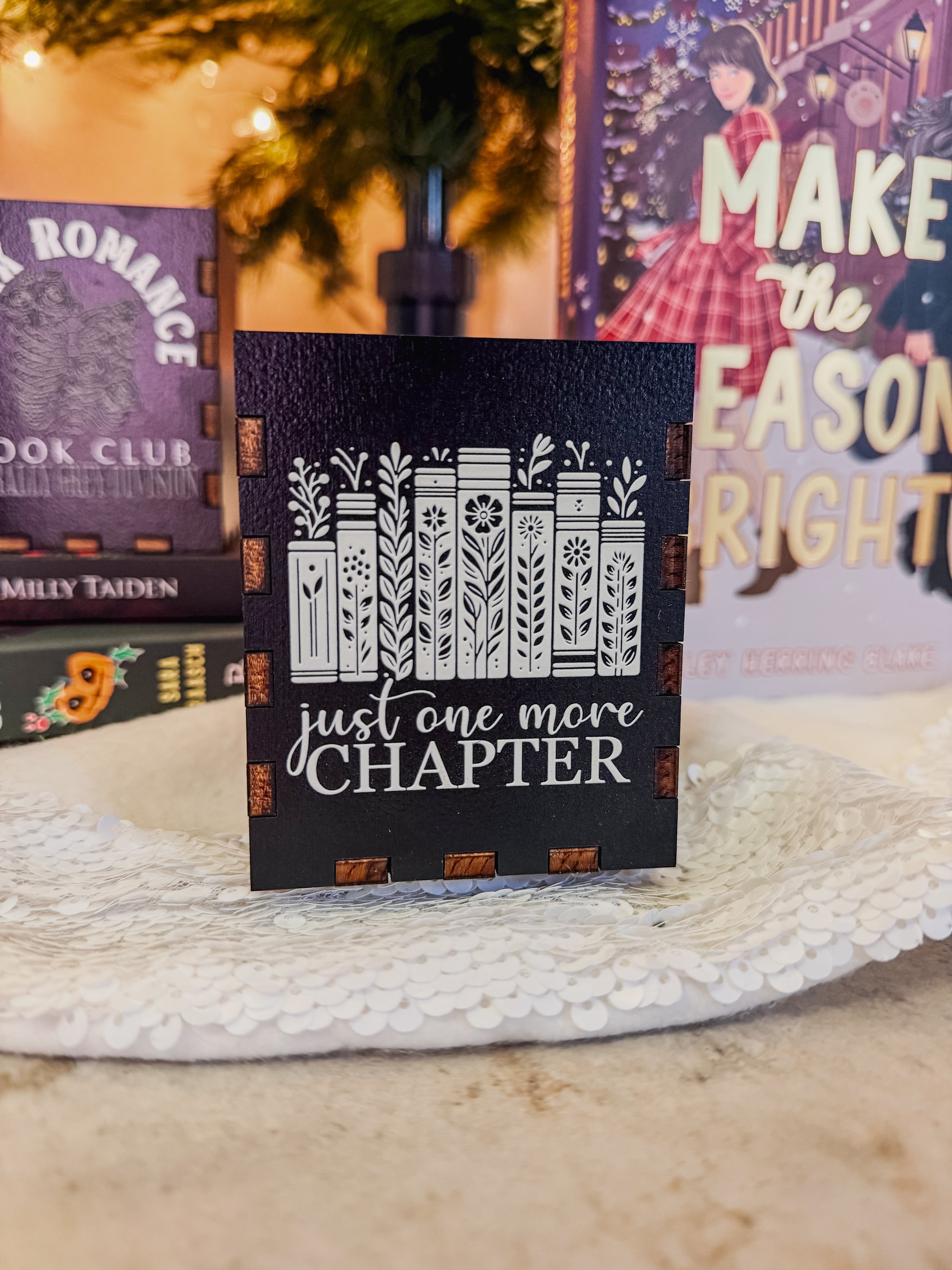 Black box with printed design with 'Just one more chapter' text and a book stack with flowers, against a physical books and Christmas tree background.