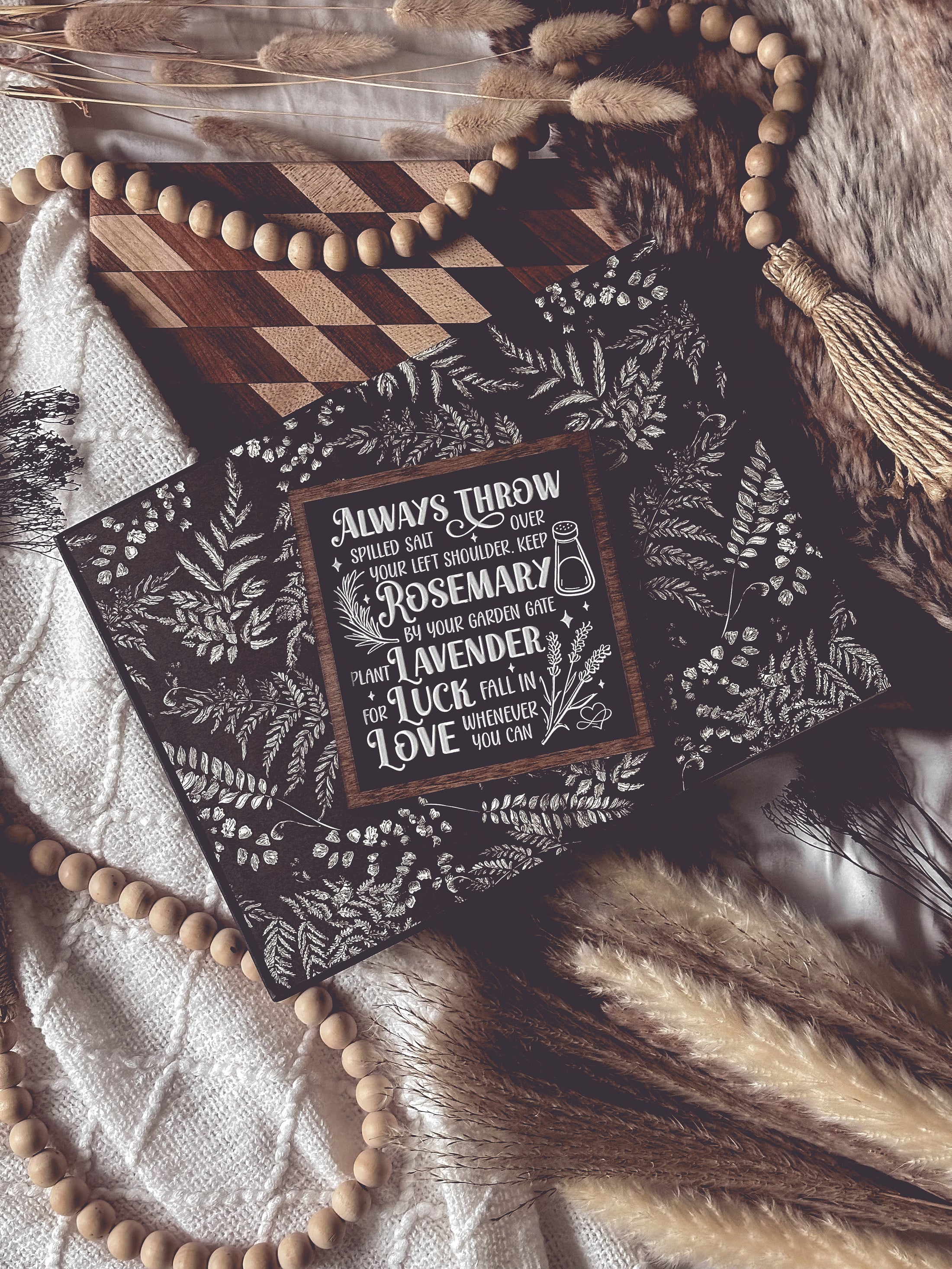 Black shelf sign with white text surrounded by a dark wood border. Graphics of a vial, lavender, rosemary and stars surround text that reads: “Always throw salt over your left shoulder. Keep rosemary by your garden gate. Plant lavender for luck. Fall in love whenever you can.” 