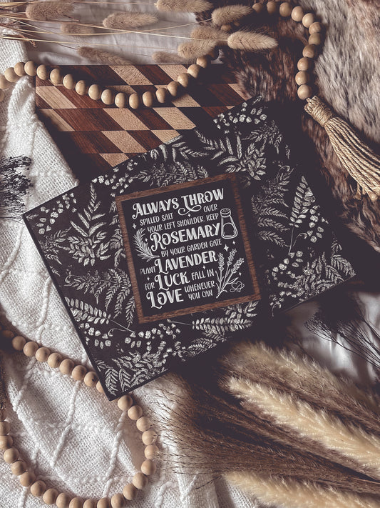 Black shelf sign with white text surrounded by a dark wood border. Graphics of a vial, lavender, rosemary and stars surround text that reads: “Always throw salt over your left shoulder. Keep rosemary by your garden gate. Plant lavender for luck. Fall in love whenever you can.” 
