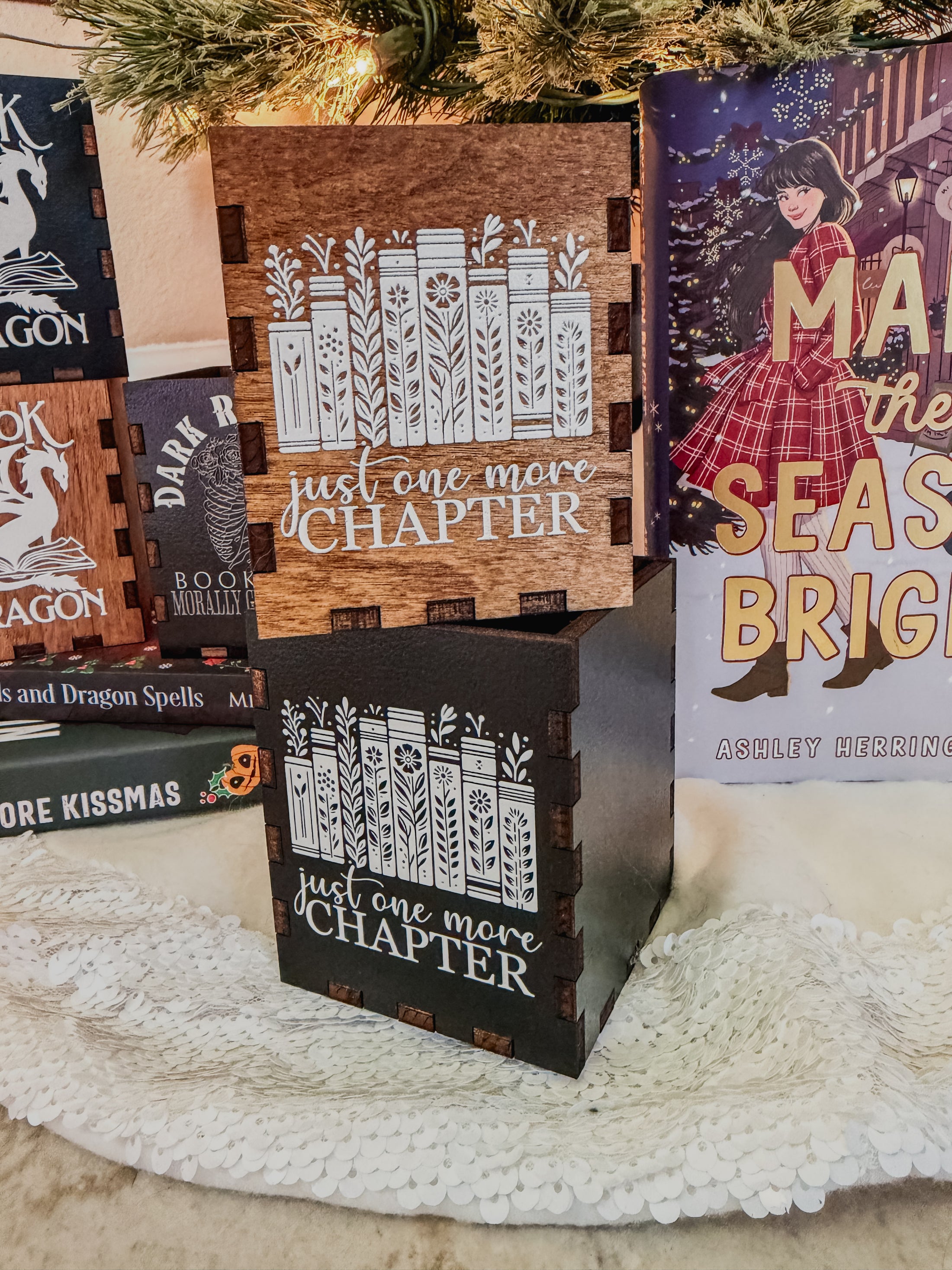 Wooden and black boxes with printed design with 'Just one more chapter' text and a book stack with flowers, against a physical books and Christmas tree background.