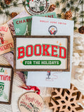 Load image into Gallery viewer, Decorative sign with a wooden frame that reads 'Booked for the Holidays' in red on a white background. © FireDrake Artistry 2025.
