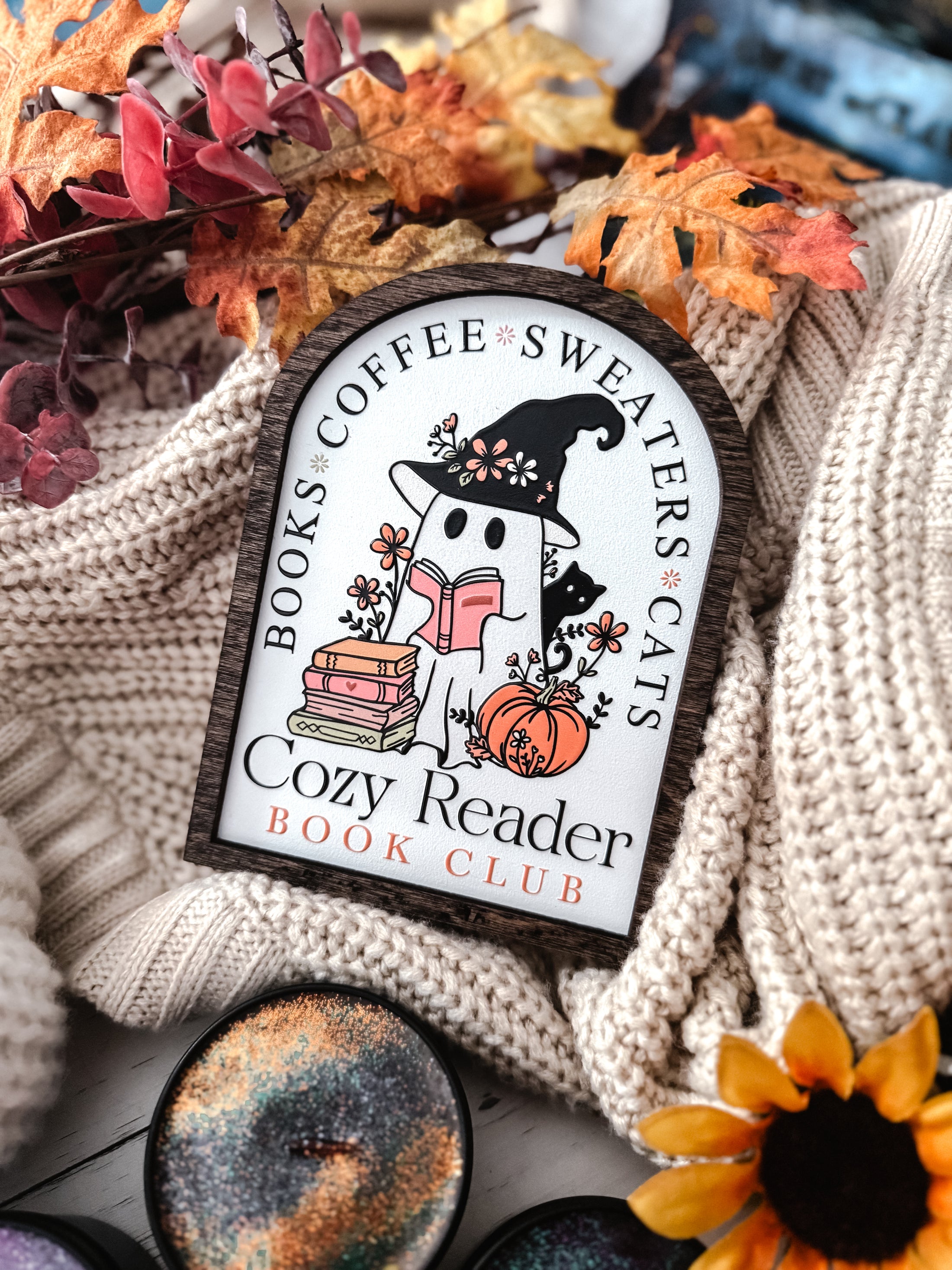 Cozy Reader Ghosty Sign. White background with a wood border with a colorful UV printed design. 
Featuring a ghost reading a book and wearing a witch hat, surrounded by flowers and a pumpkin. Text above reads “Books. Coffee. Sweaters. Cats.” While the text below reads “Cozy Reader Book Club.” 