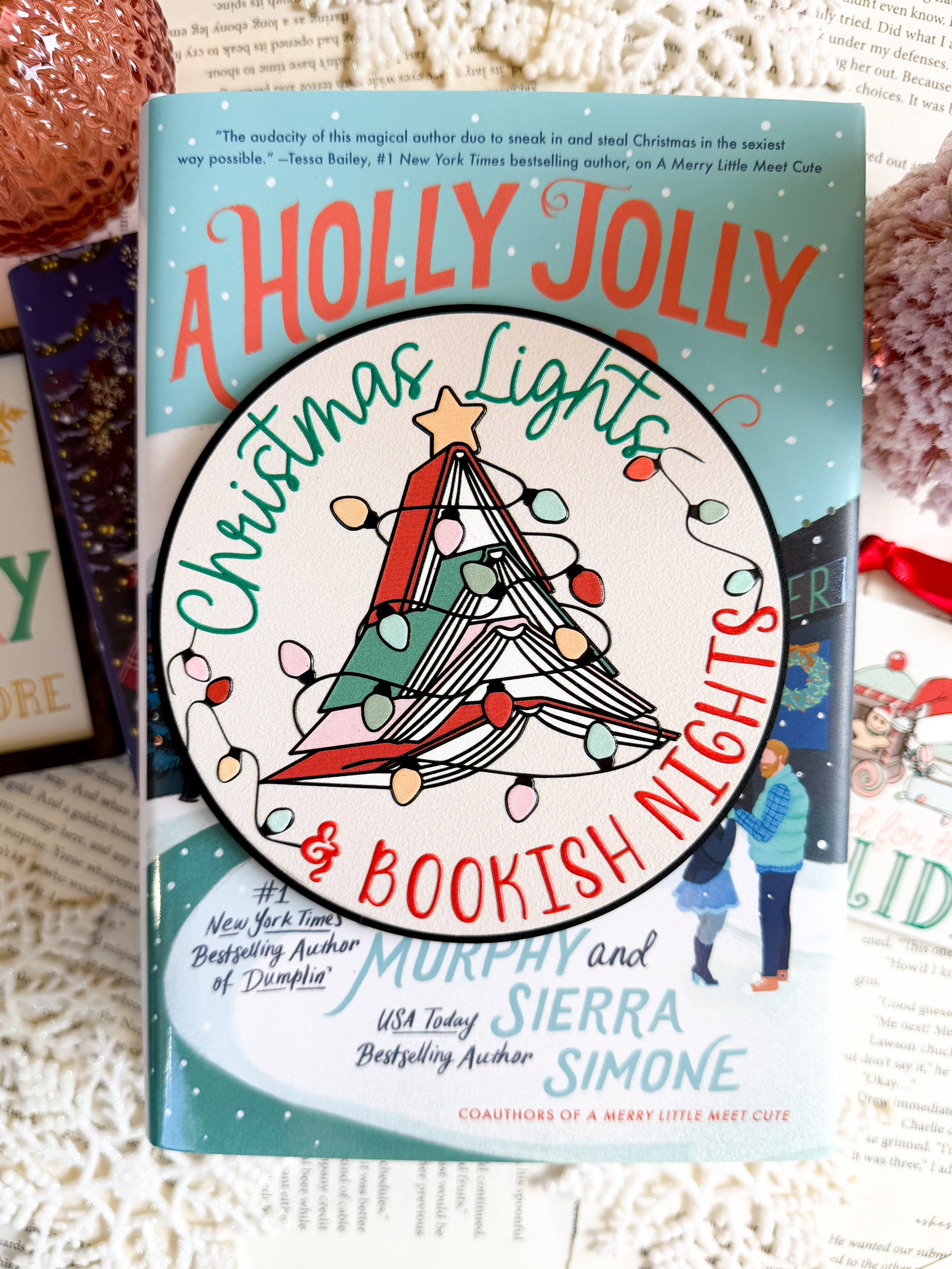 Round shelf sign with a white background. Image of books stacked into a Christmas tree surrounded by lights. The surrounding text reads "Christmas Lights" above in green, with "& Bookish Nights" below in red. © FireDrake Artistry 2025