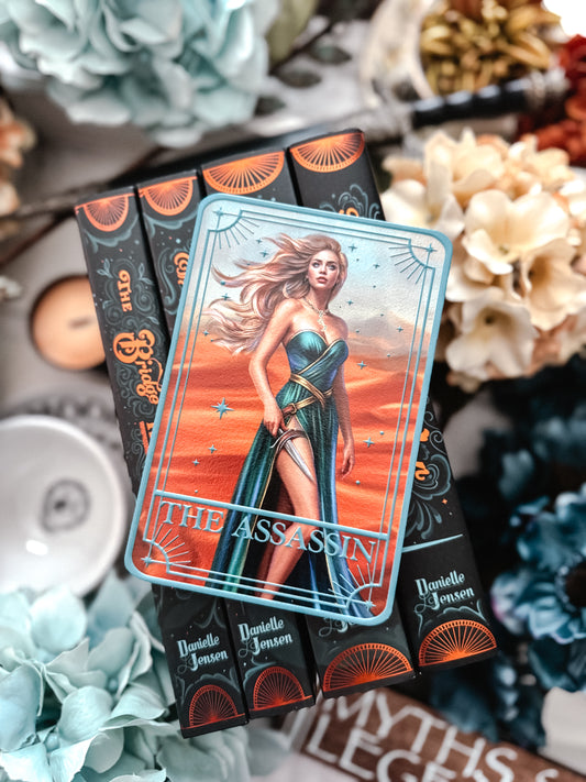 Tarot card titled 'The Assassin' on top of a book stack with flowers and decorative items in the background. 