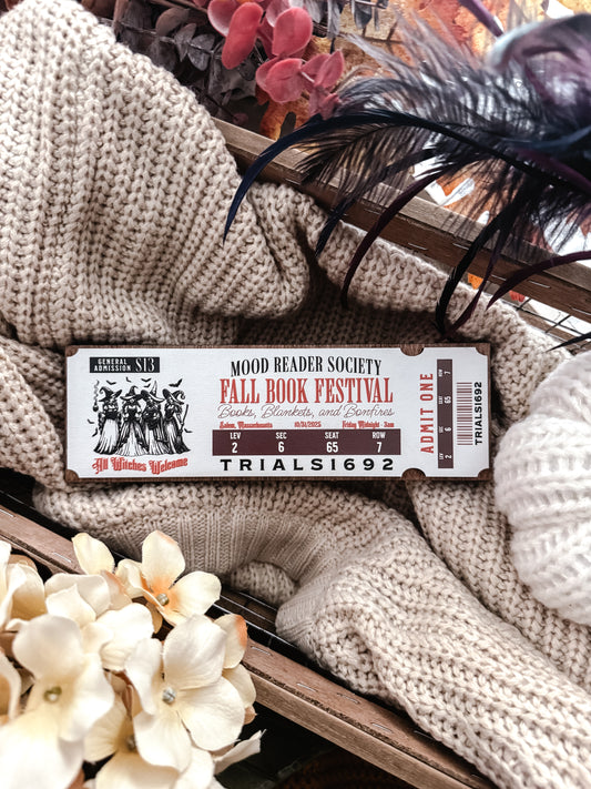 Fall Book Festival Ticket Sign. Thick Wooden sign with a white background shaped like a ticket. Orange text that reads “Fall Book Festival” with a date and ticket details. Featuring an image of witches, which beneath reads “witches welcome”. ©️FireDrake Artistry 2025