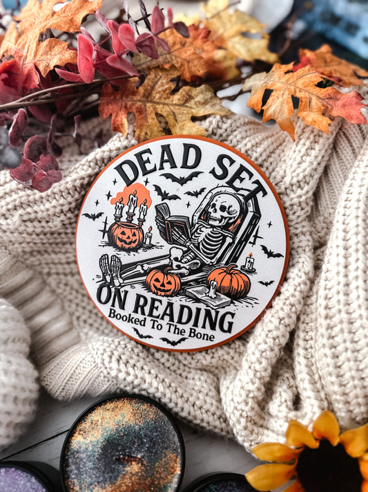 Dead Set on Reading Sign - Orange. White round sign with a skeleton lounging and reading a book surrounded by pumpkins. 