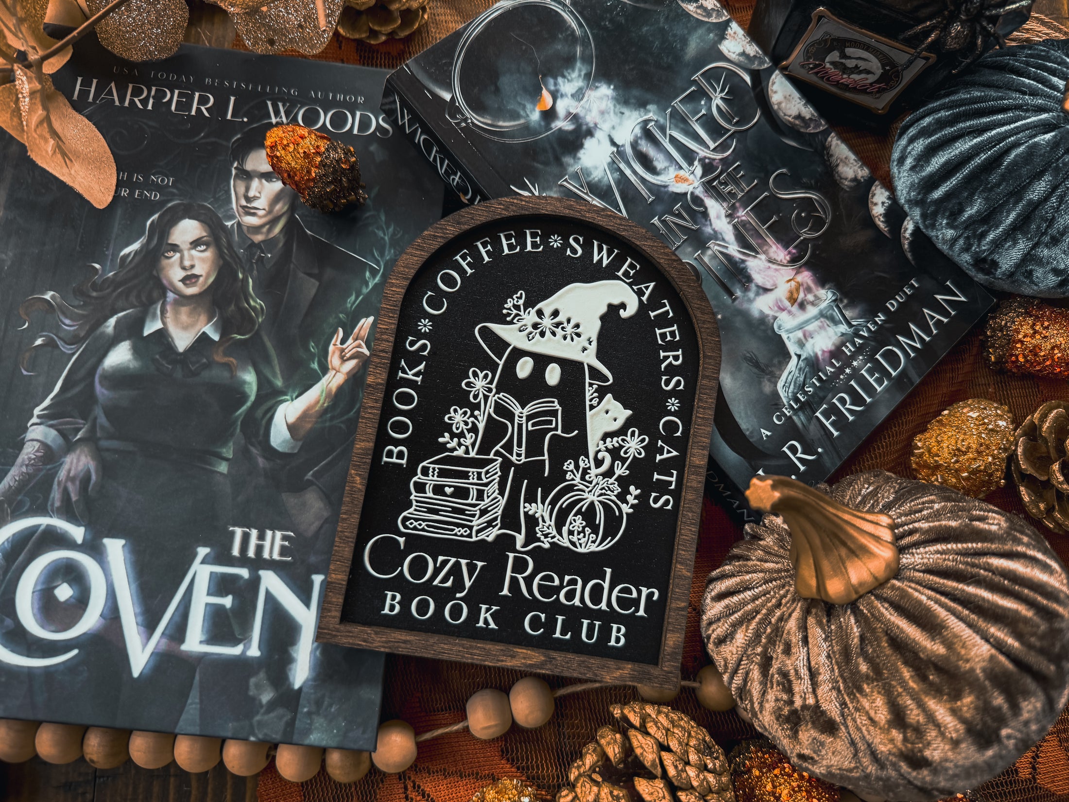 Cozy Reader Ghosty Sign. Black background with a dark wood border with a white design engraved. 
Featuring a ghost reading a book and wearing a witch hat, surrounded by flowers and a pumpkin. Text above reads “Books. Coffee. Sweaters. Cats.” While the text below reads “Cozy Reader Book Club.” 